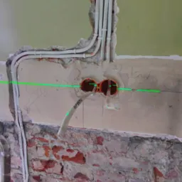 Dwa pomarańczowe otwory pod gniazdka w ścianie z widoczną zieloną linią lasera, prowadzone białe kable w peszlach, ściana z fragmentami cegły i resztkami tynku.