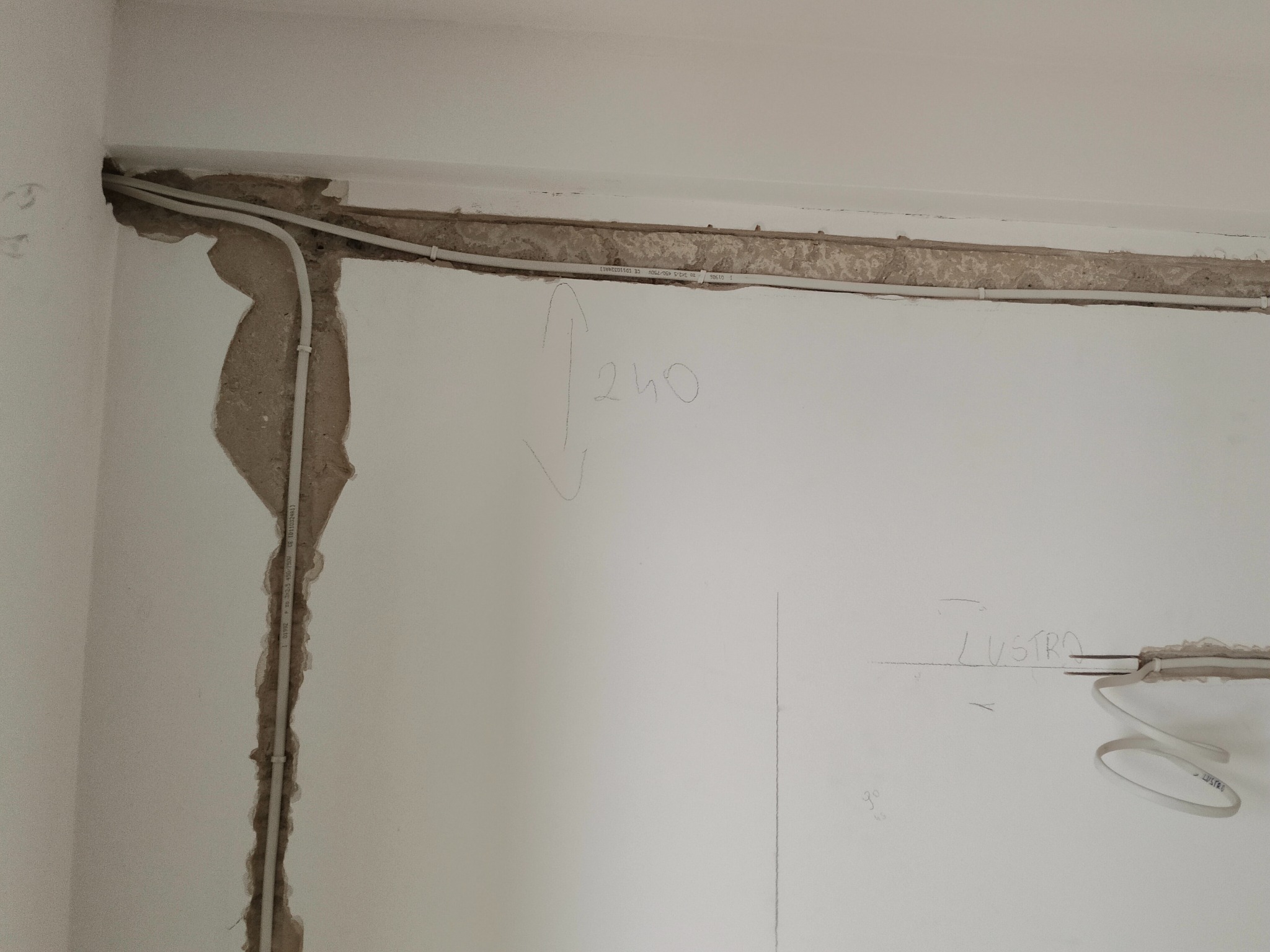 Białe przewody elektryczne prowadzone w bruzdach w ścianie podczas remontu. Widoczne odręczne notatki na ścianie, w tym wymiar '240' i słowo 'LUSTRO'.