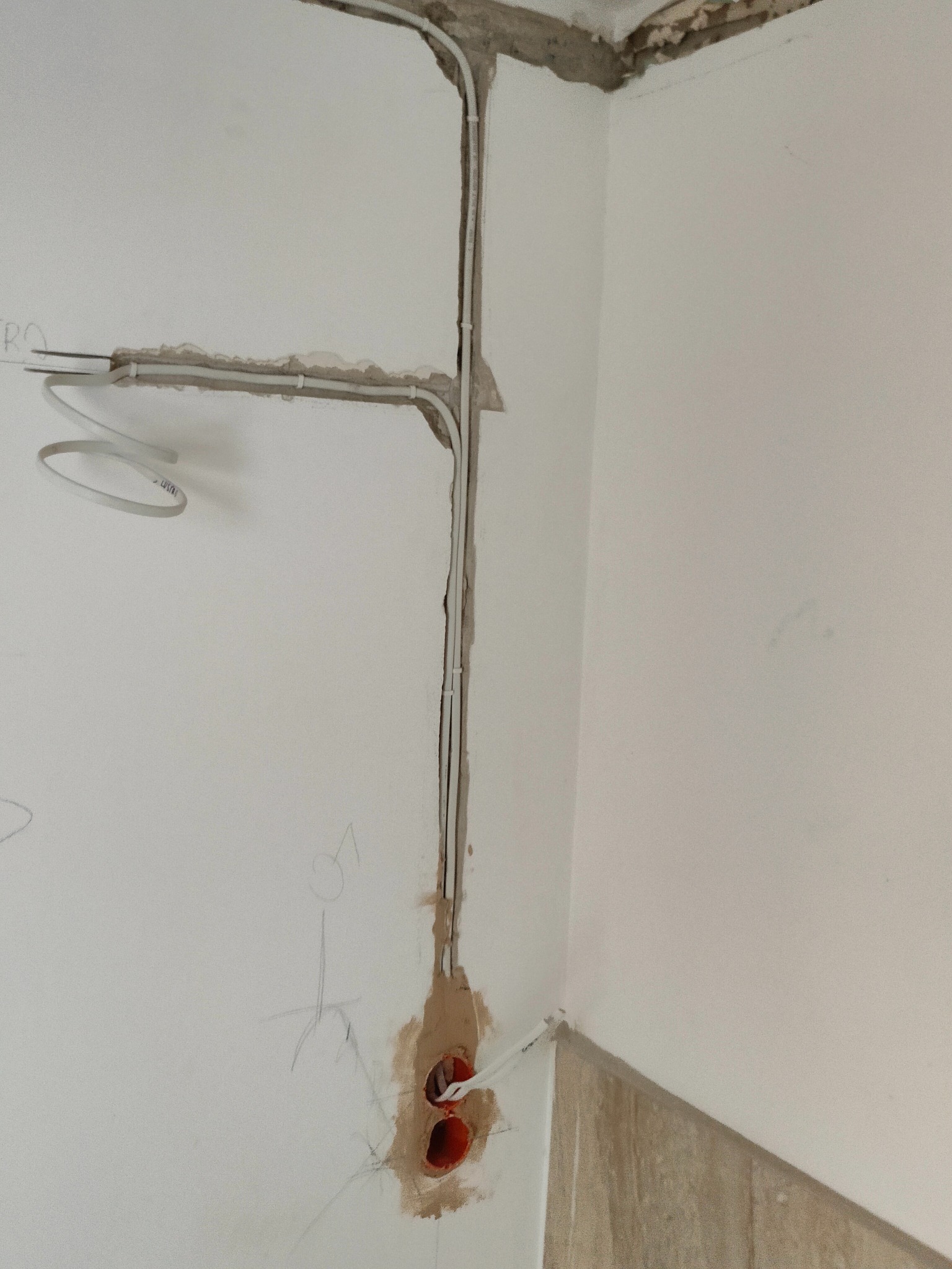 Białe przewody elektryczne w bruzdach na ścianie, widoczne puszki instalacyjne z wystającymi kablami, surowy stan wykończenia.