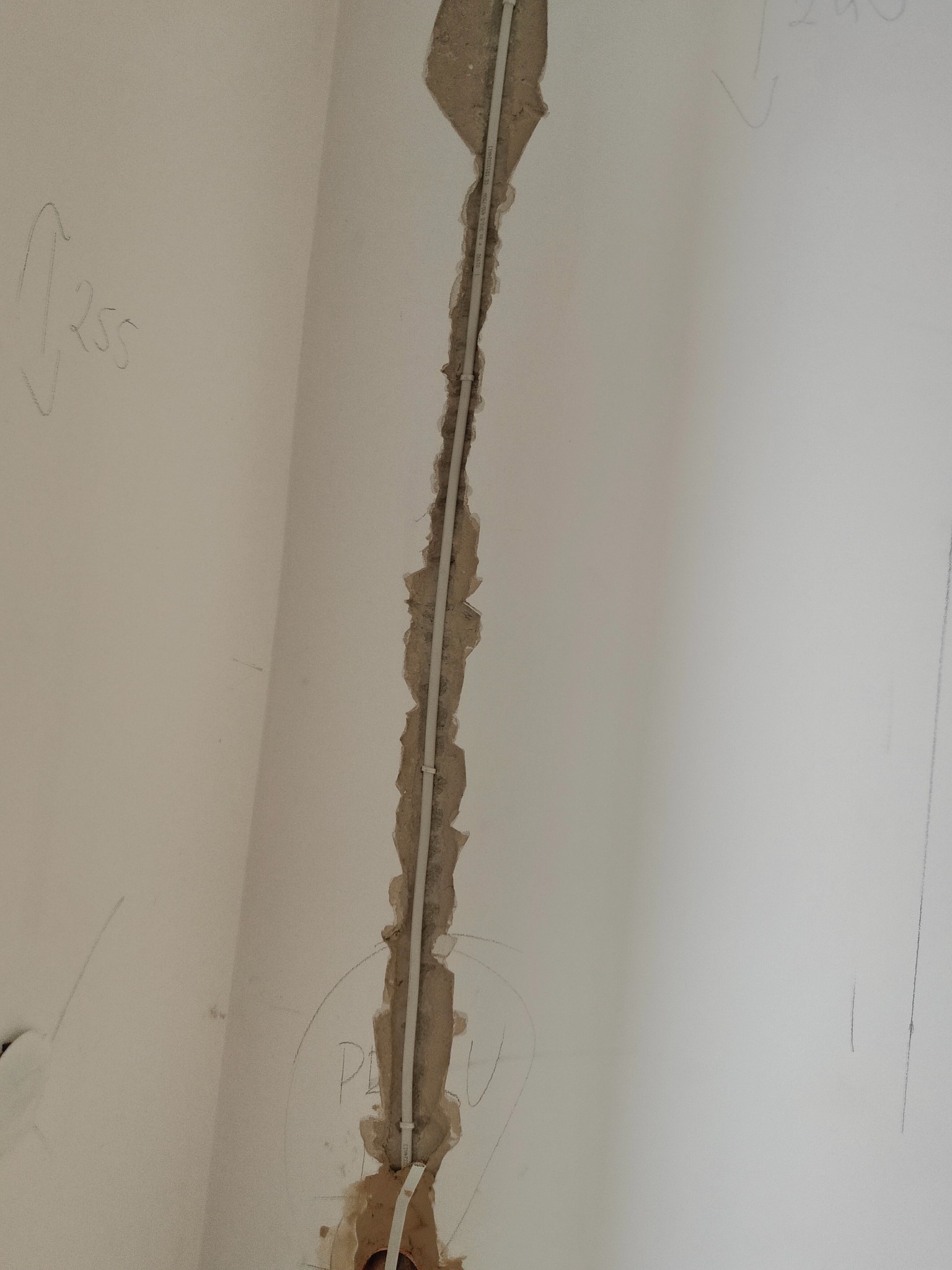 Biała rurka instalacyjna poprowadzona w bruździe w ścianie, widoczne oznaczenia ołówkiem na ścianie, surowy stan przed wykończeniem.