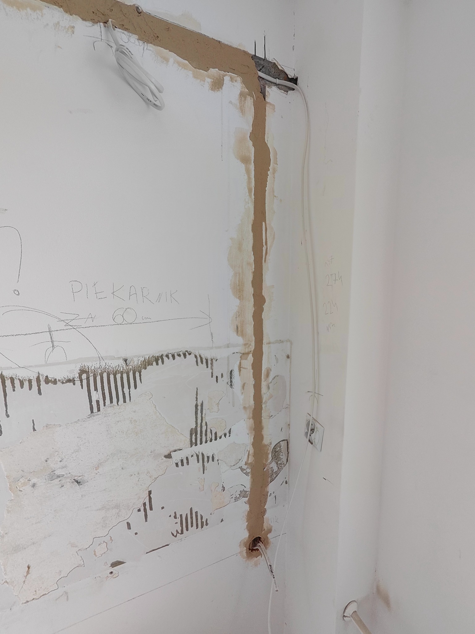 Biała ściana w trakcie remontu z wyżłobionymi bruzdami pod kable elektryczne, widoczne przewody, gniazdko, resztki tapety i odręczne notatki ołówkiem, w tym napis 'PIEKARNIK za 60cm'.