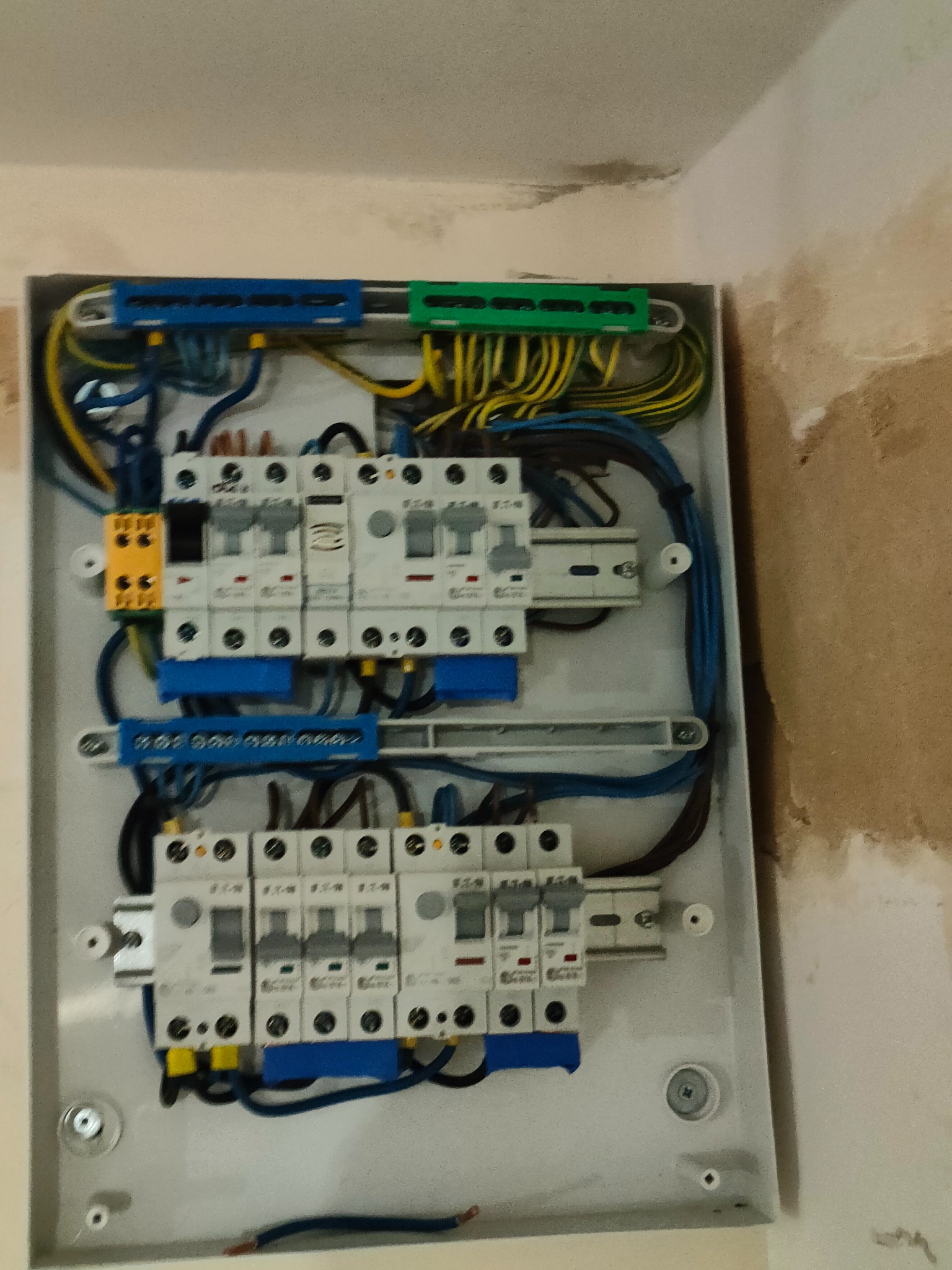 Rozdzielnica elektryczna z zamontowanymi bezpiecznikami i podłączonymi przewodami, widoczne kable w kolorach niebieskim, żółto-zielonym i czarnym, skrzynka w kolorze białym, surowa ściana w tle.