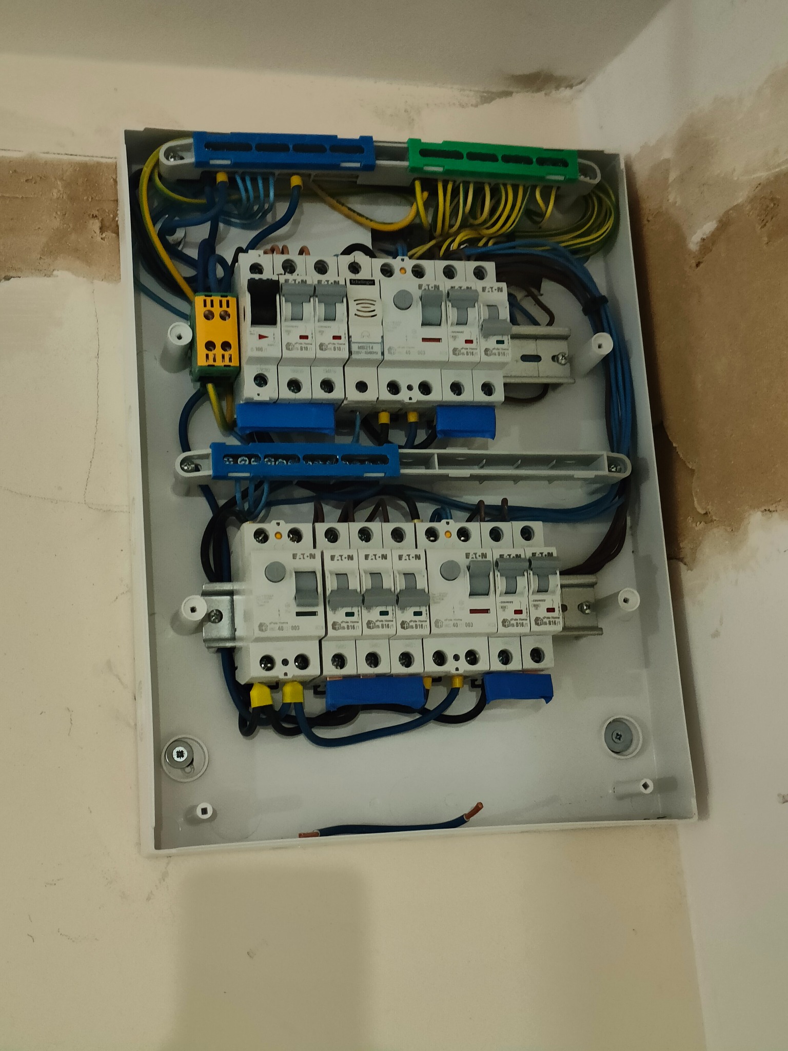 Rozdzielnica elektryczna Eaton z zainstalowanymi bezpiecznikami, widoczne kable w kolorach niebieskim, żółto-zielonym i czarnym, zamontowana na ścianie w trakcie prac wykończeniowych.