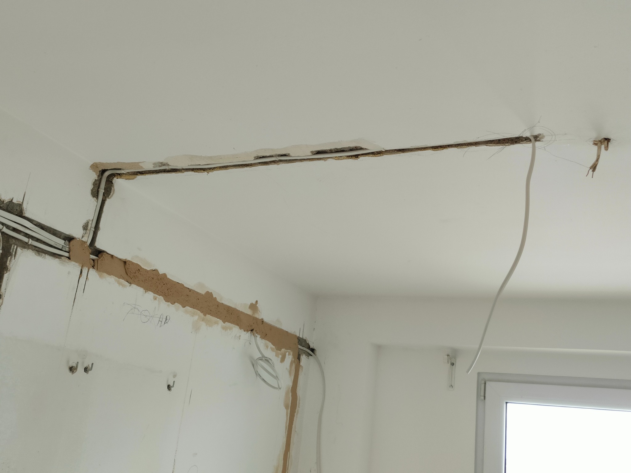 Wykute bruzdy w ścianie i suficie z poprowadzonymi przewodami elektrycznymi, widoczne ślady po zaprawie, fragment okna w tle.