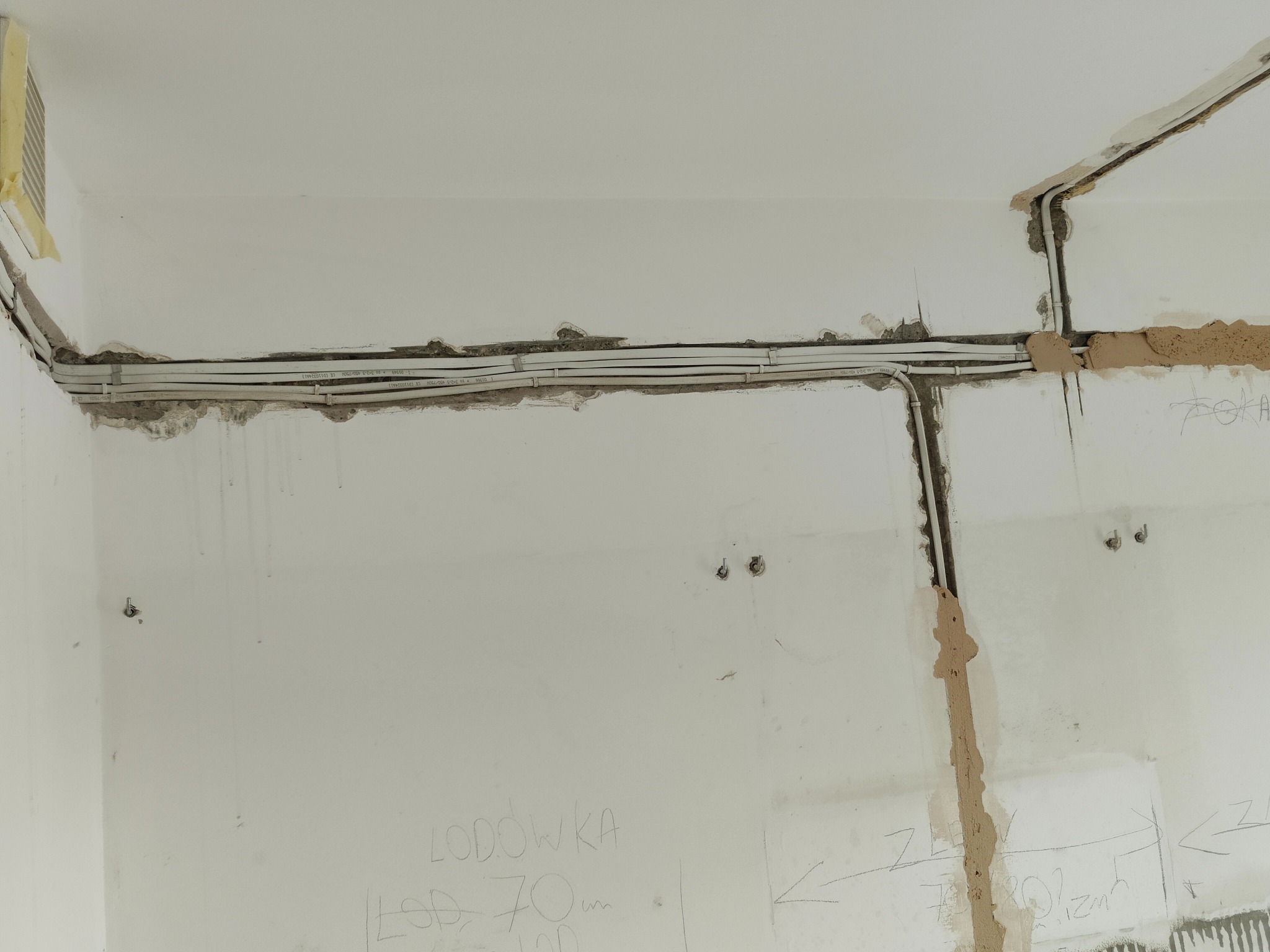 Białe przewody elektryczne zamocowane do ściany z wyżłobionymi bruzdami, częściowo zamaskowane gipsem, z widocznymi notatkami ołówkiem na ścianie.