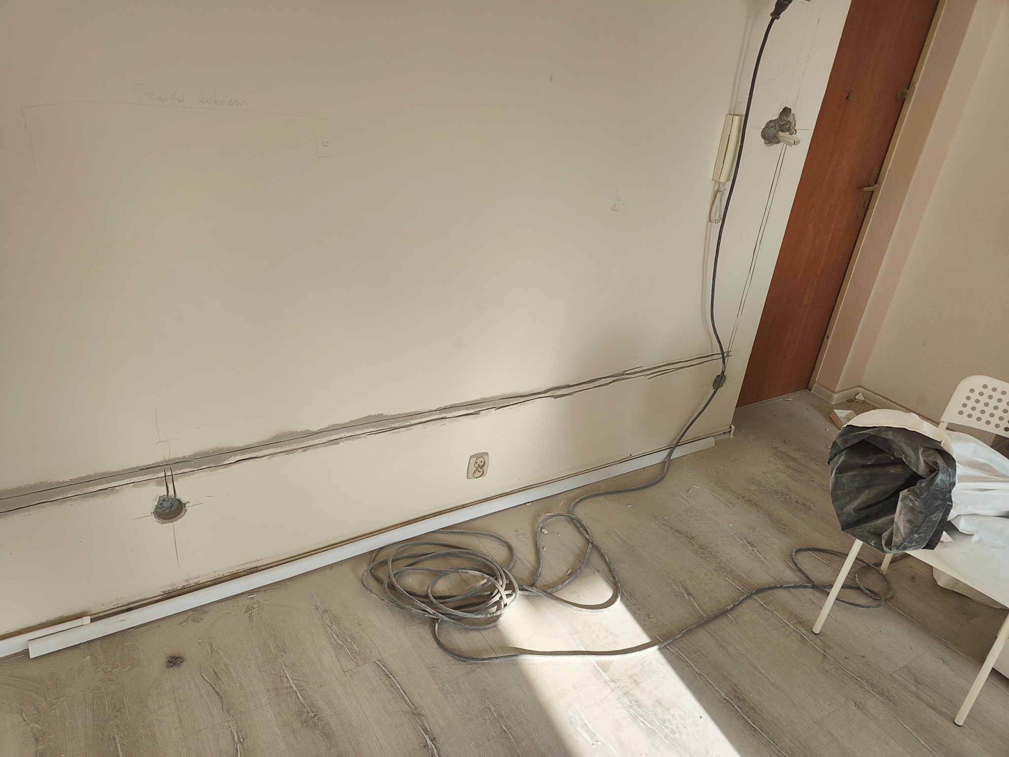Ściana z wyciętymi bruzdami pod przewody, widoczne kable na podłodze, gniazdko elektryczne, instalacja w trakcie wykonywania, ślady po wierceniu, napis ołówkiem na ścianie.