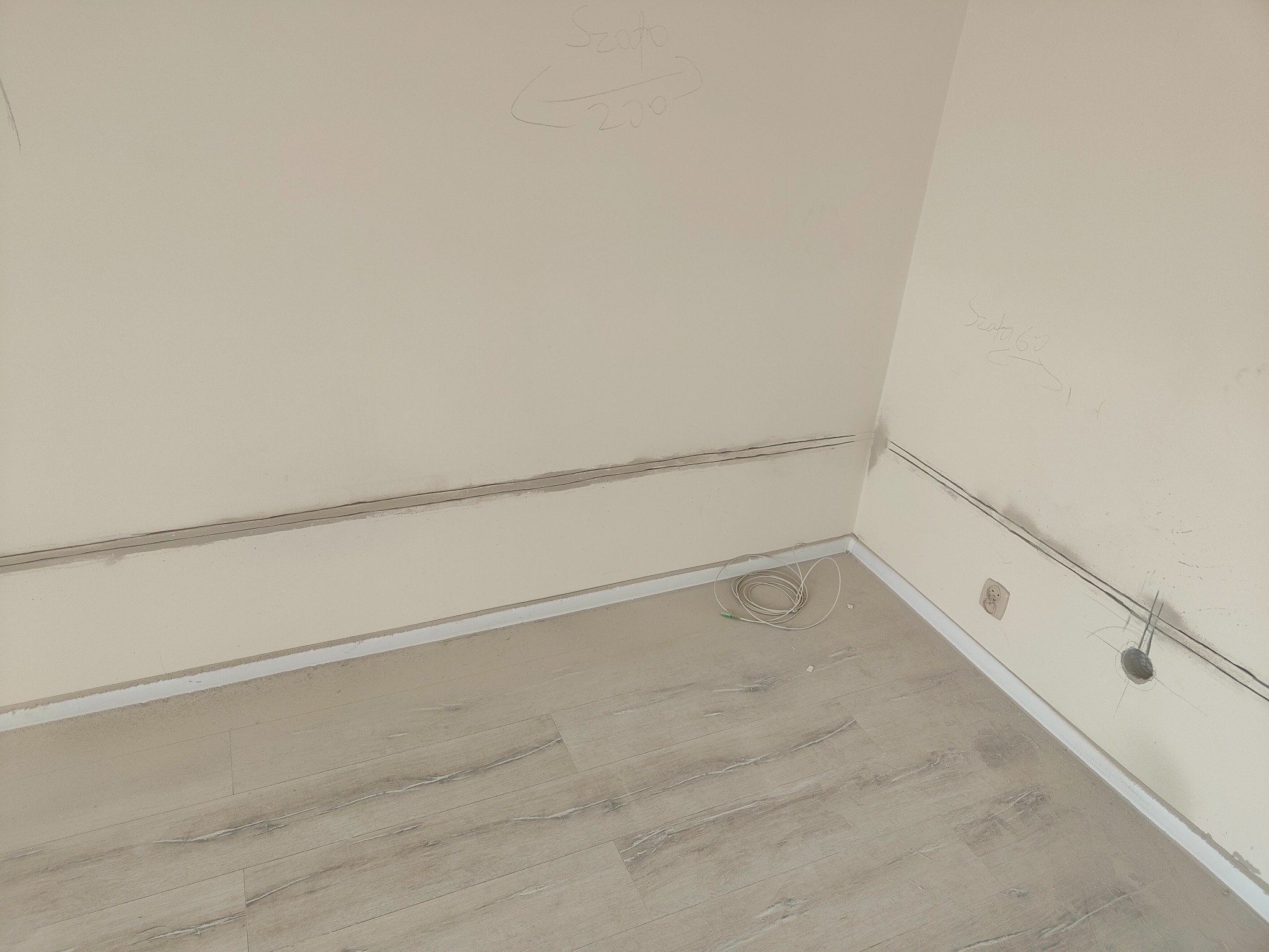 Przygotowanie do rozprowadzenia przewodów elektrycznych w nowym budynku, widoczne bruzdy w ścianach, gniazdko, otwór na puszkę oraz zwój kabli na podłodze z paneli.
