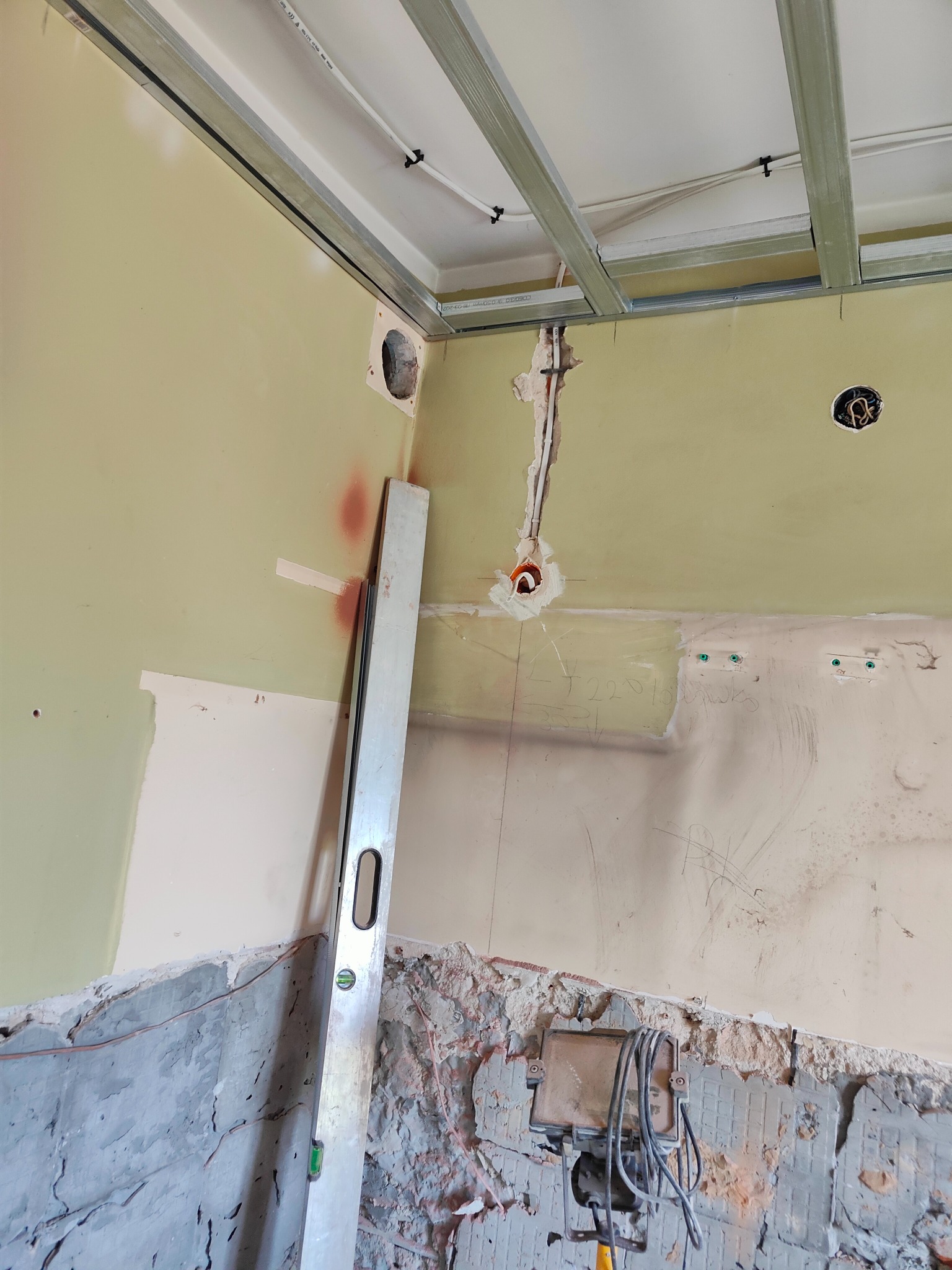 Wnętrze w trakcie remontu z widocznymi kablami elektrycznymi wystającymi ze ściany, poziomica oparta o ścianę, metalowa konstrukcja sufitu podwieszanego, oraz lampa robocza na dole.