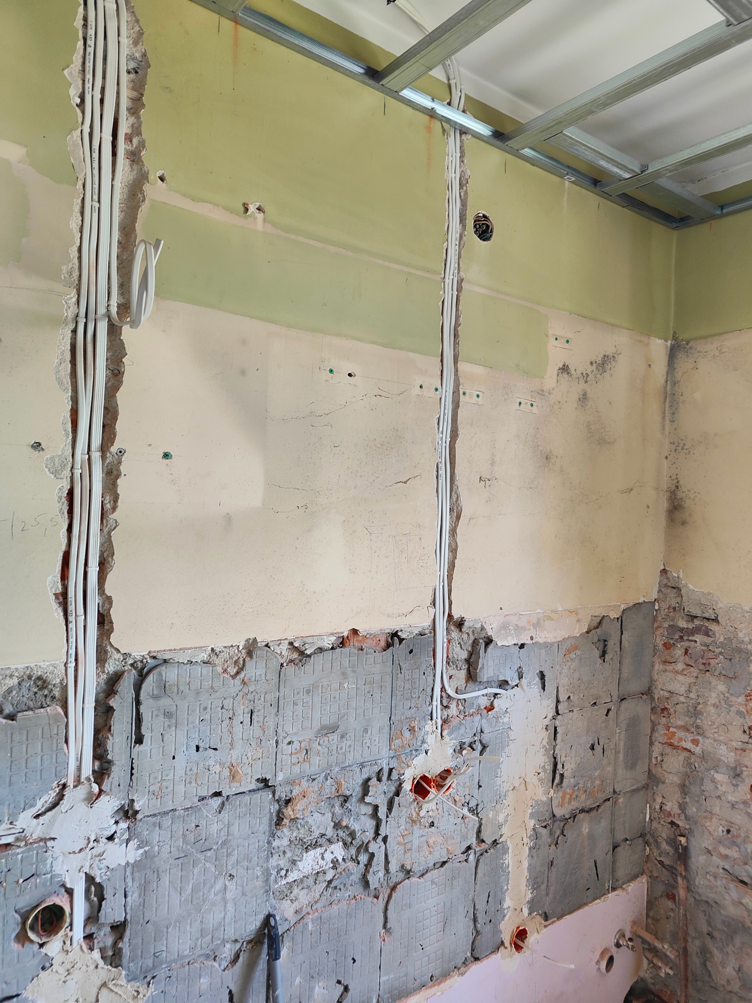 Ściana w trakcie remontu z bruzdami pod kable elektryczne i puszkami instalacyjnymi, widoczne fragmenty starej instalacji i konstrukcja sufitu podwieszanego.