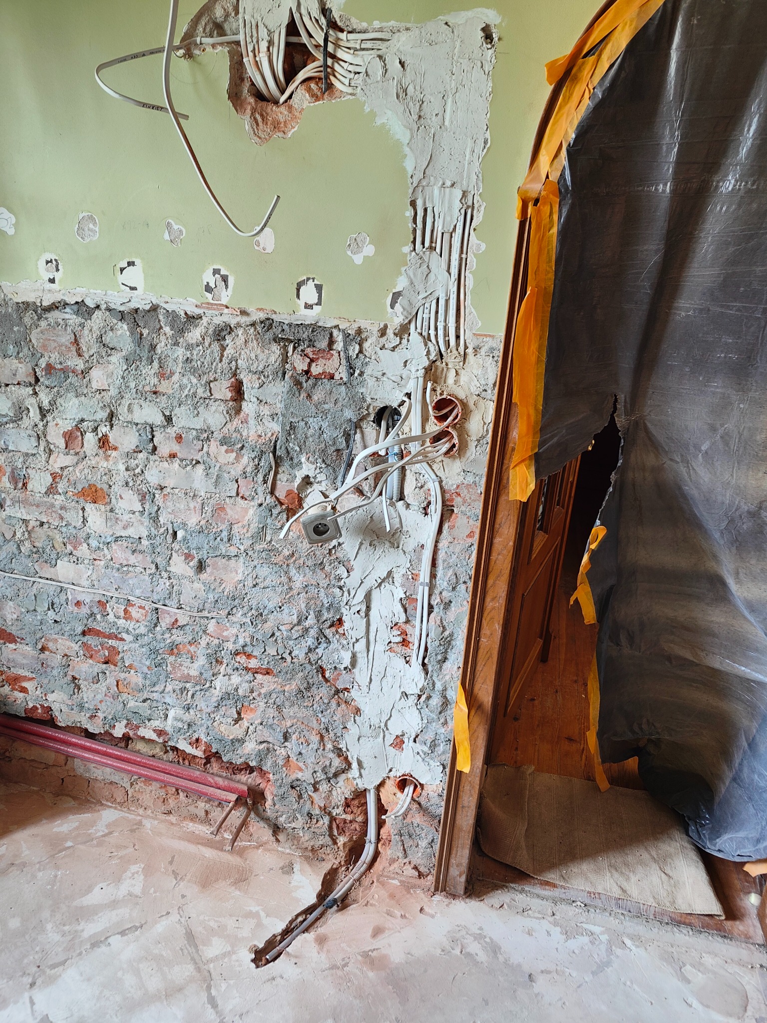 Ściana w trakcie remontu z odsłoniętą instalacją elektryczną i fragmentem muru ceglanego, zabezpieczenie folią malarską przy framudze drzwi.