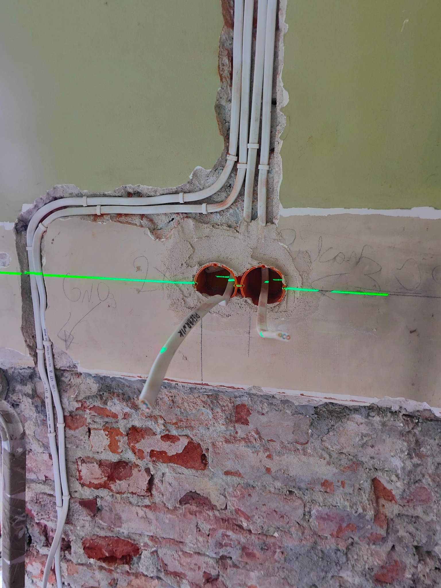 Dwa pomarańczowe otwory pod gniazdka w ścianie z widoczną zieloną linią lasera, prowadzone białe kable w peszlach, ściana z fragmentami cegły i resztkami tynku.
