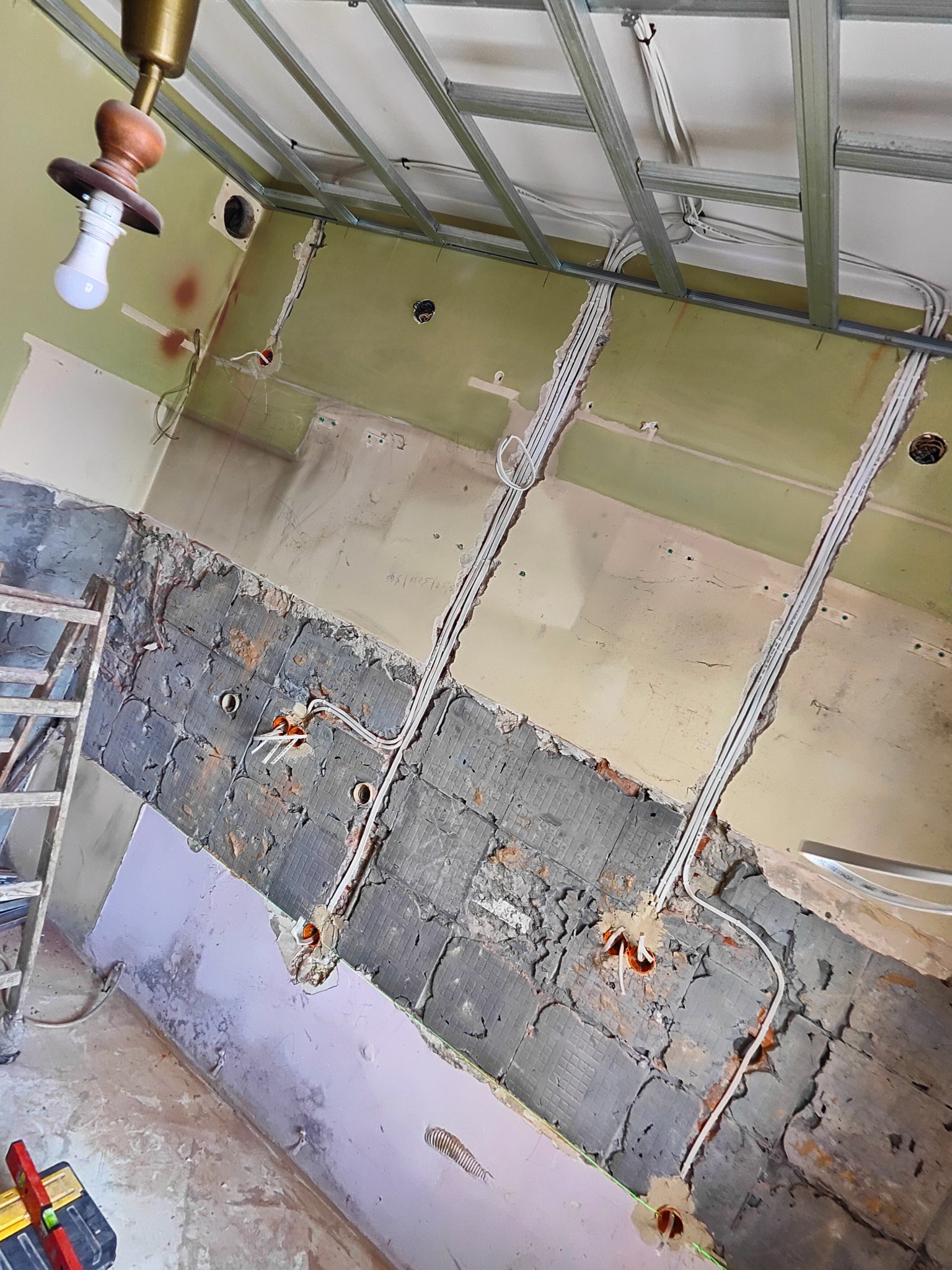 Wnętrze w trakcie remontu z widocznymi przewodami elektrycznymi w ścianach, zamontowanym stelażem sufitowym, drabina i skrzynka z narzędziami w rogu.