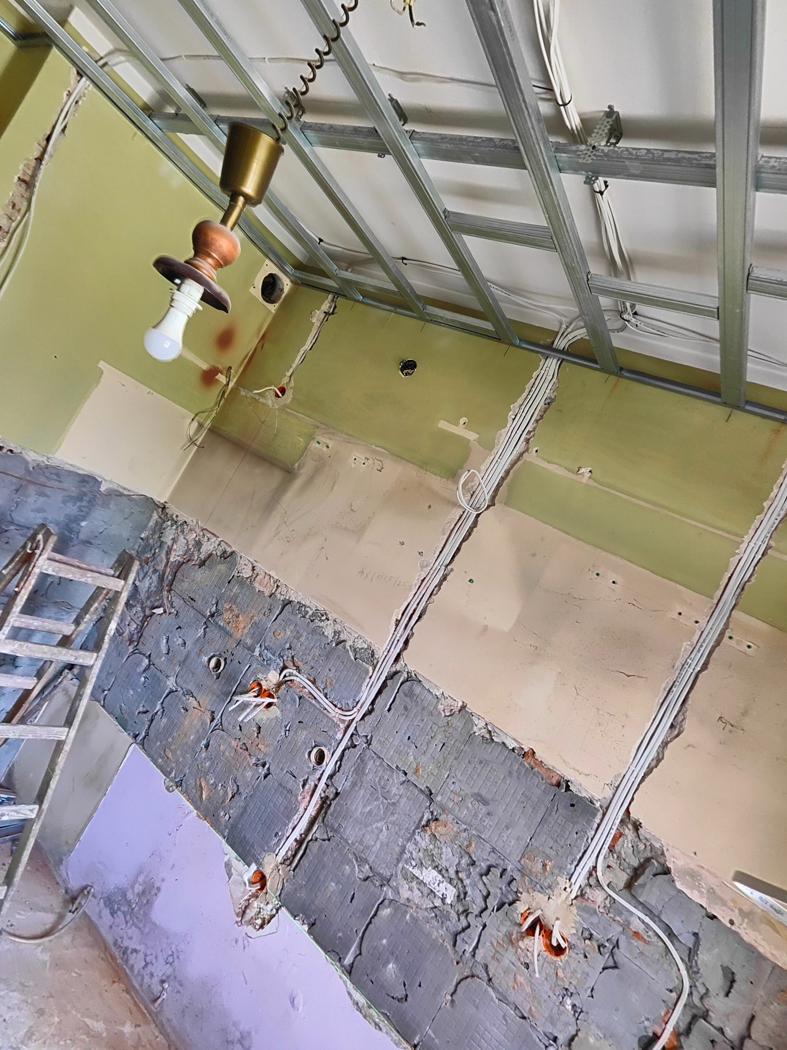 Wnętrze w trakcie remontu z widoczną instalacją elektryczną: przewody w ścianach, puszki instalacyjne, metalowy stelaż sufitu podwieszanego i wisząca lampa tymczasowa.