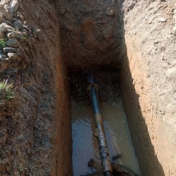 MM INSTAL-WIERT Marcin Twaróg - Wykop z wodą i rurą w ziemi, widok z góry. Ściany wykopu z ziemi, na górze kamienie i trawa. Rura częściowo zanurzona w wodzie.