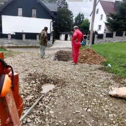 MM INSTAL-WIERT Marcin Twaróg - Dwóch mężczyzn nad wykopem na żwirowym podłożu, w tle domy jednorodzinne. Widoczny fragment maszyny z pomarańczowymi słuchawkami ochronnymi.