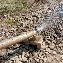MM INSTAL-WIERT Marcin Twaróg - Woda tryskająca z metalowej rury na kamienistym podłożu, prawdopodobnie podczas prac związanych z instalacją podziemną.