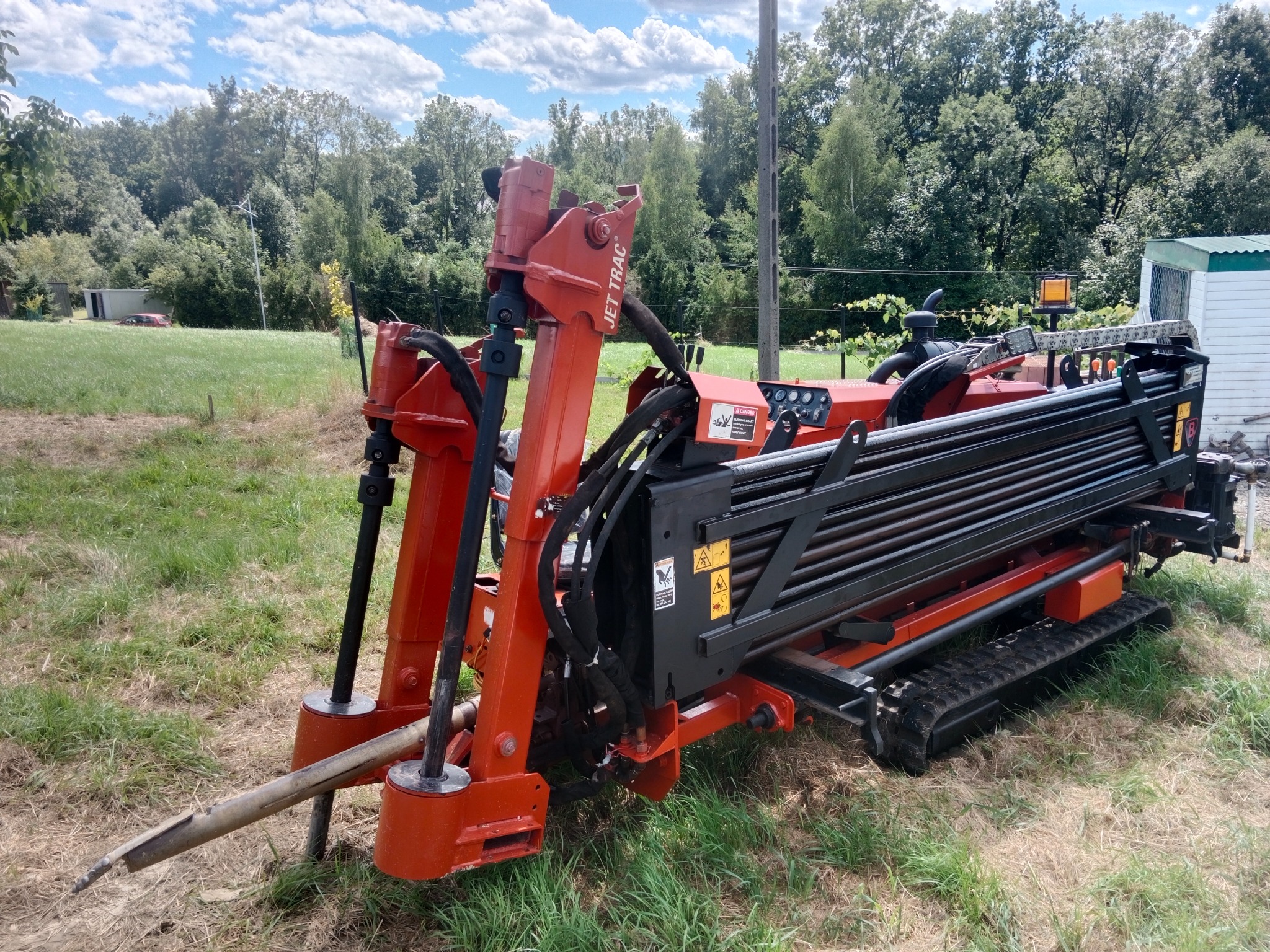Pomarańczowa wiertnica Jet Trac na gąsienicach, gotowa do pracy na trawiastym terenie z drzewami w tle. Widoczne przewody hydrauliczne i elementy sterujące.