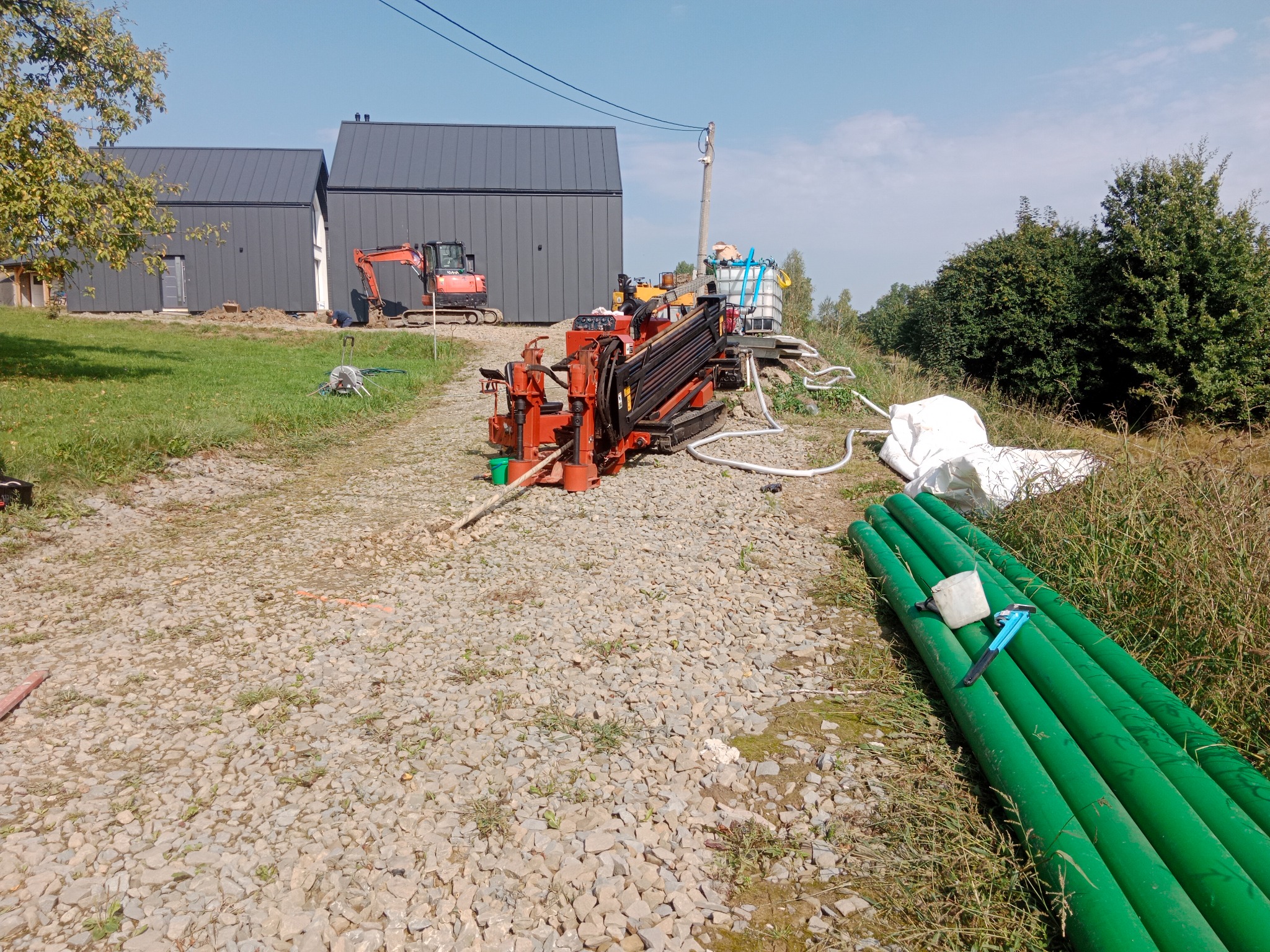 Pomarańczowa wiertnica pozioma na żwirowej drodze, obok zielone rury i biały zbiornik. W tle domy z szarej blachy i mała koparka.