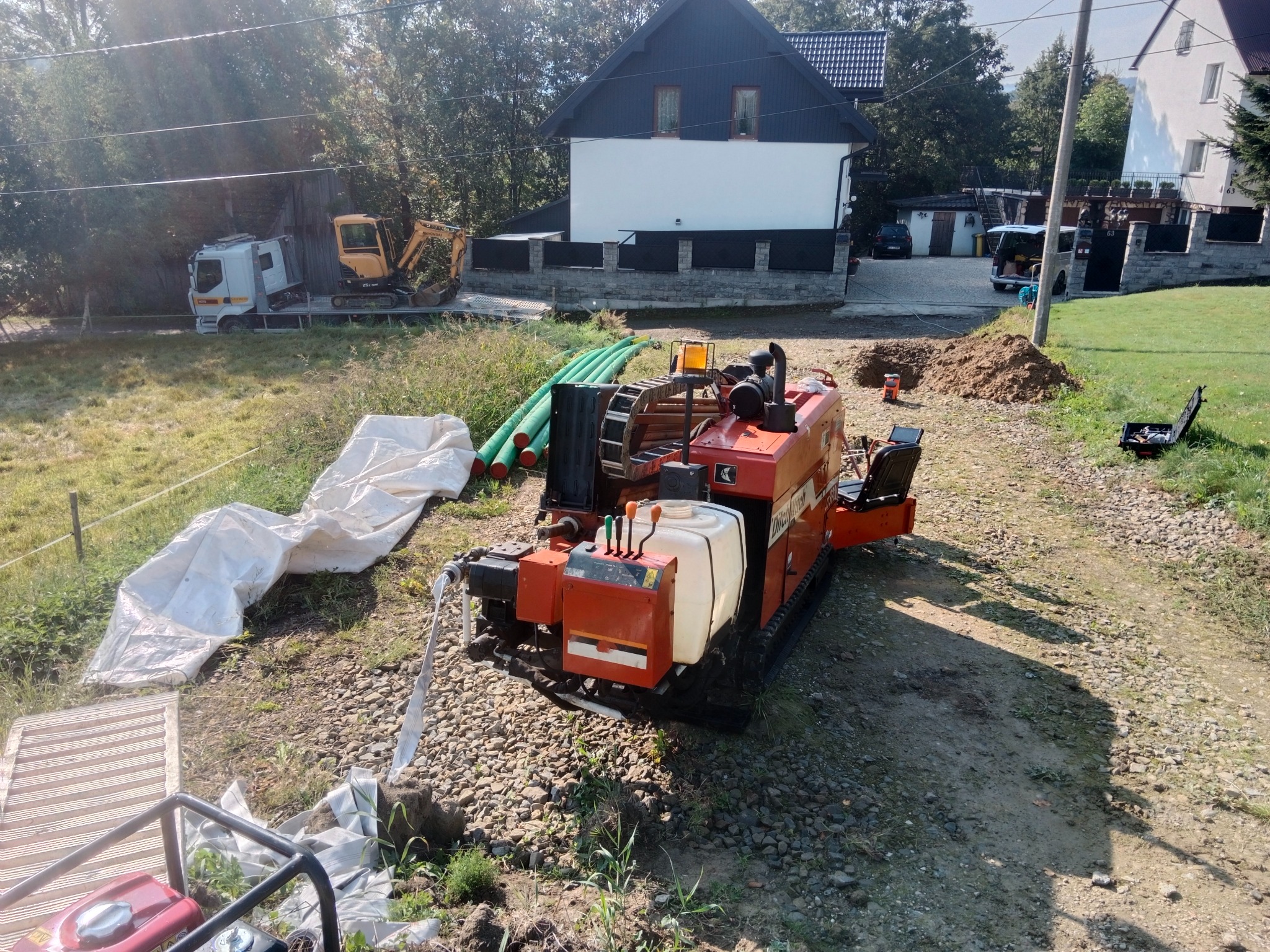 Pomarańczowa maszyna do robót ziemnych na żwirowym podłożu, z zielonymi rurami w tle i wykopem ziemnym. Widoczny dom w tle.