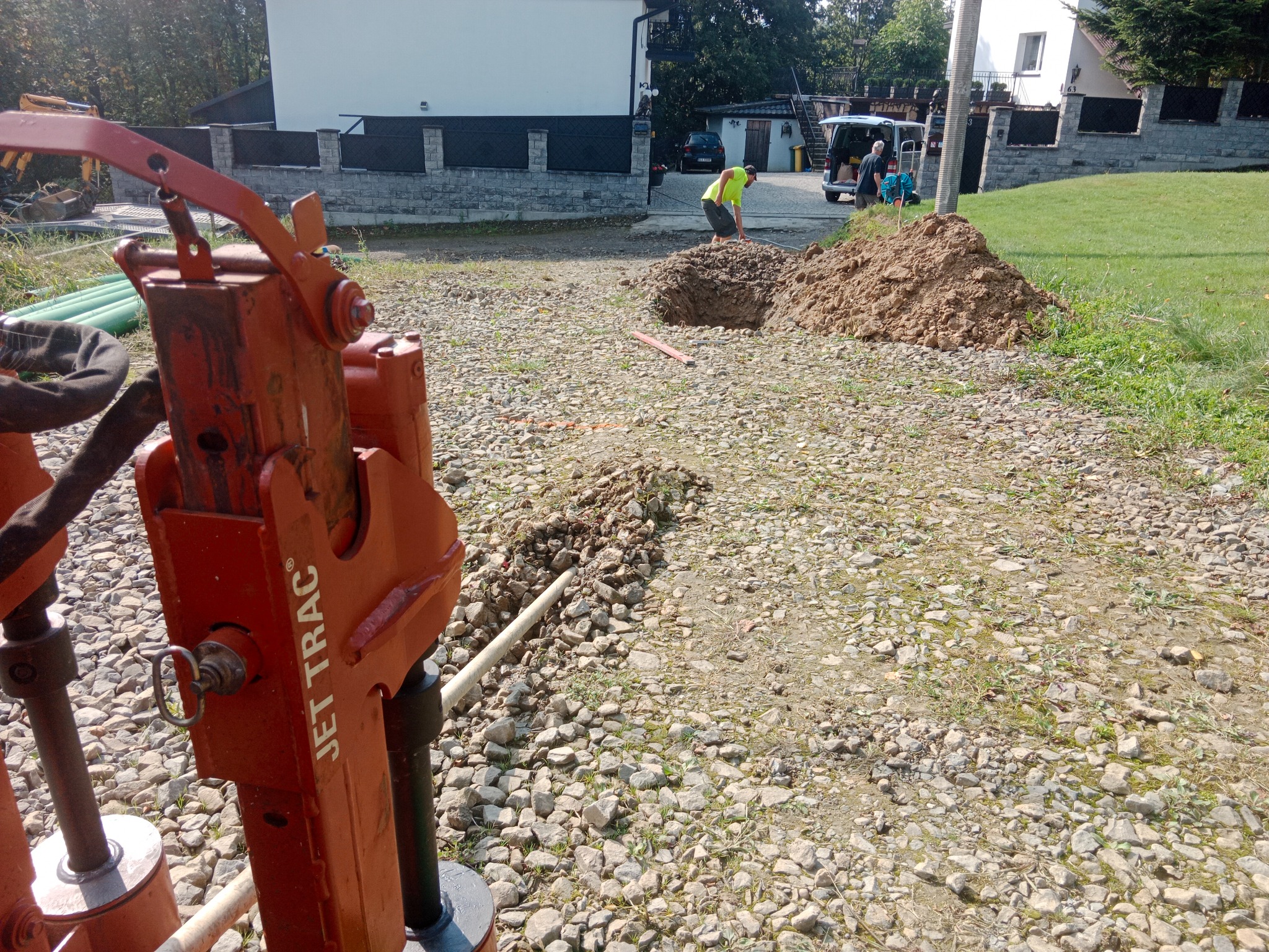 Pomarańczowa maszyna Jet Trac na żwirowym podłożu, w tle wykop i pracownik w żółtej koszulce, sugerujący prace ziemne. Widoczne budynki w tle.