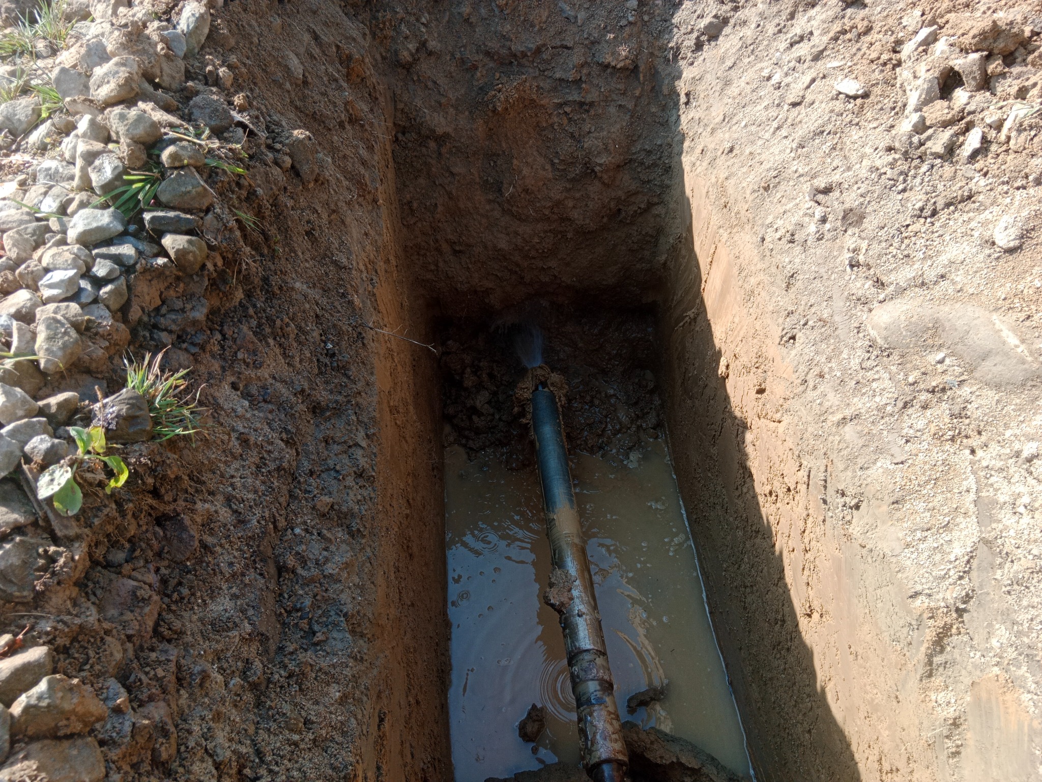 Wykop z wodą i rurą w ziemi, widok z góry. Ściany wykopu z ziemi, na górze kamienie i trawa. Rura częściowo zanurzona w wodzie.