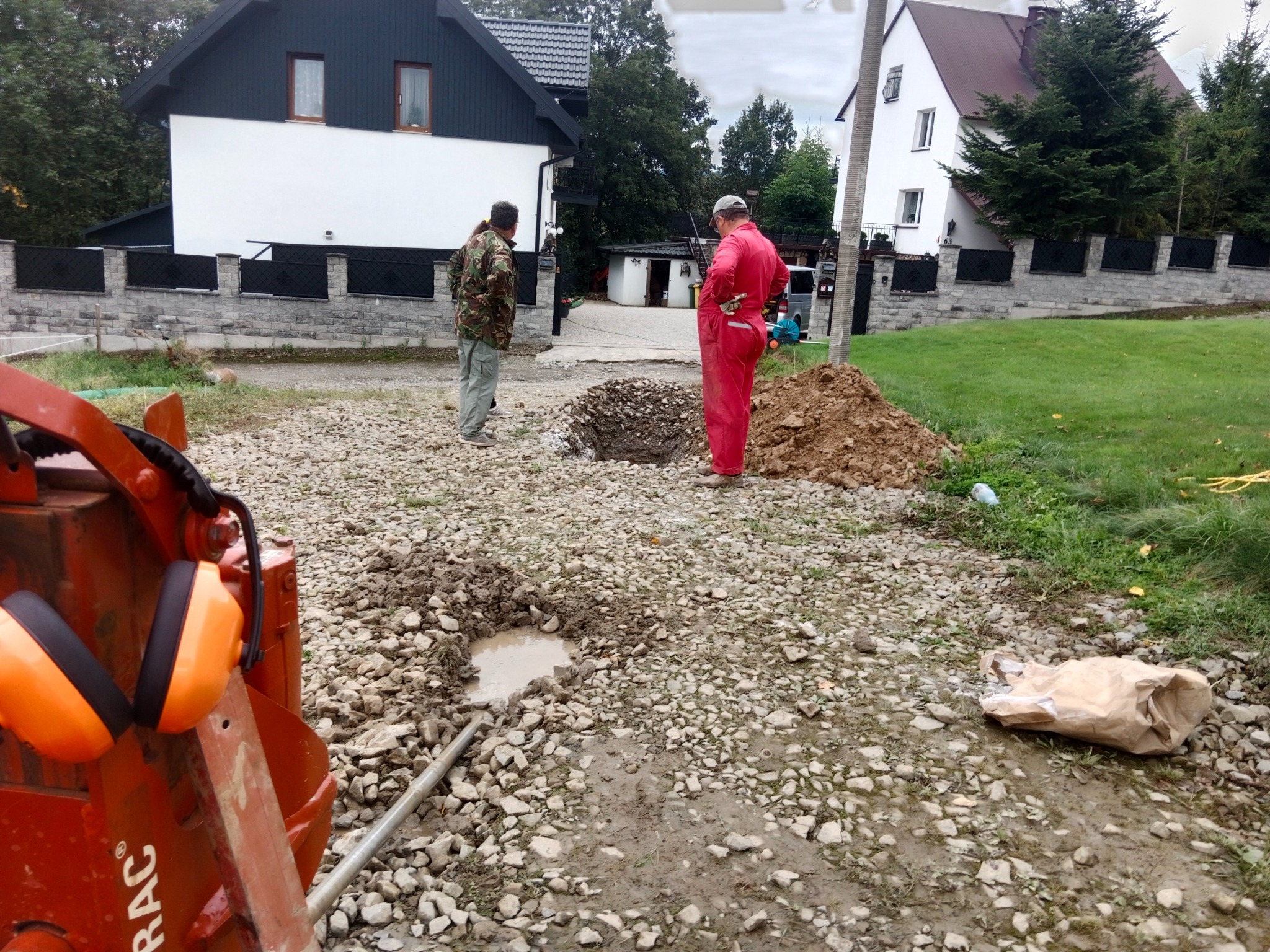 Dwóch mężczyzn nad wykopem na żwirowym podłożu, w tle domy jednorodzinne. Widoczny fragment maszyny z pomarańczowymi słuchawkami ochronnymi.