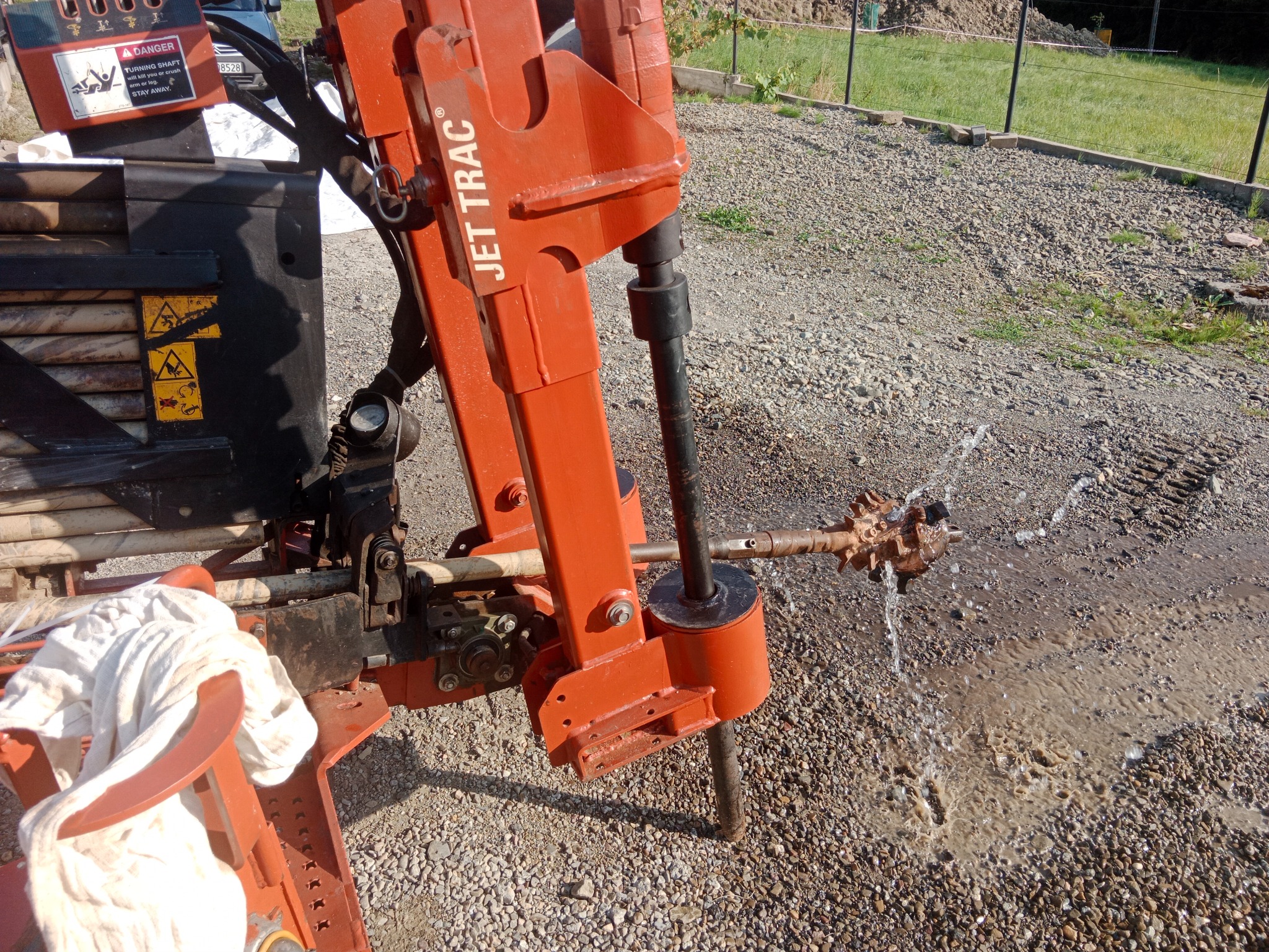 Pomarańczowa wiertnica Jet Trac w trakcie pracy, woda tryska spod wiertła. Widoczne ostrzeżenie o obracającym się wale. Tło: trawa i kamieniste podłoże.