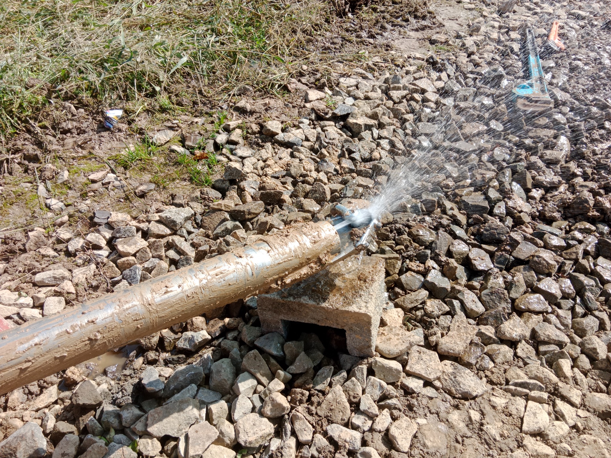 Woda tryskająca z metalowej rury na kamienistym podłożu, prawdopodobnie podczas prac związanych z instalacją podziemną.