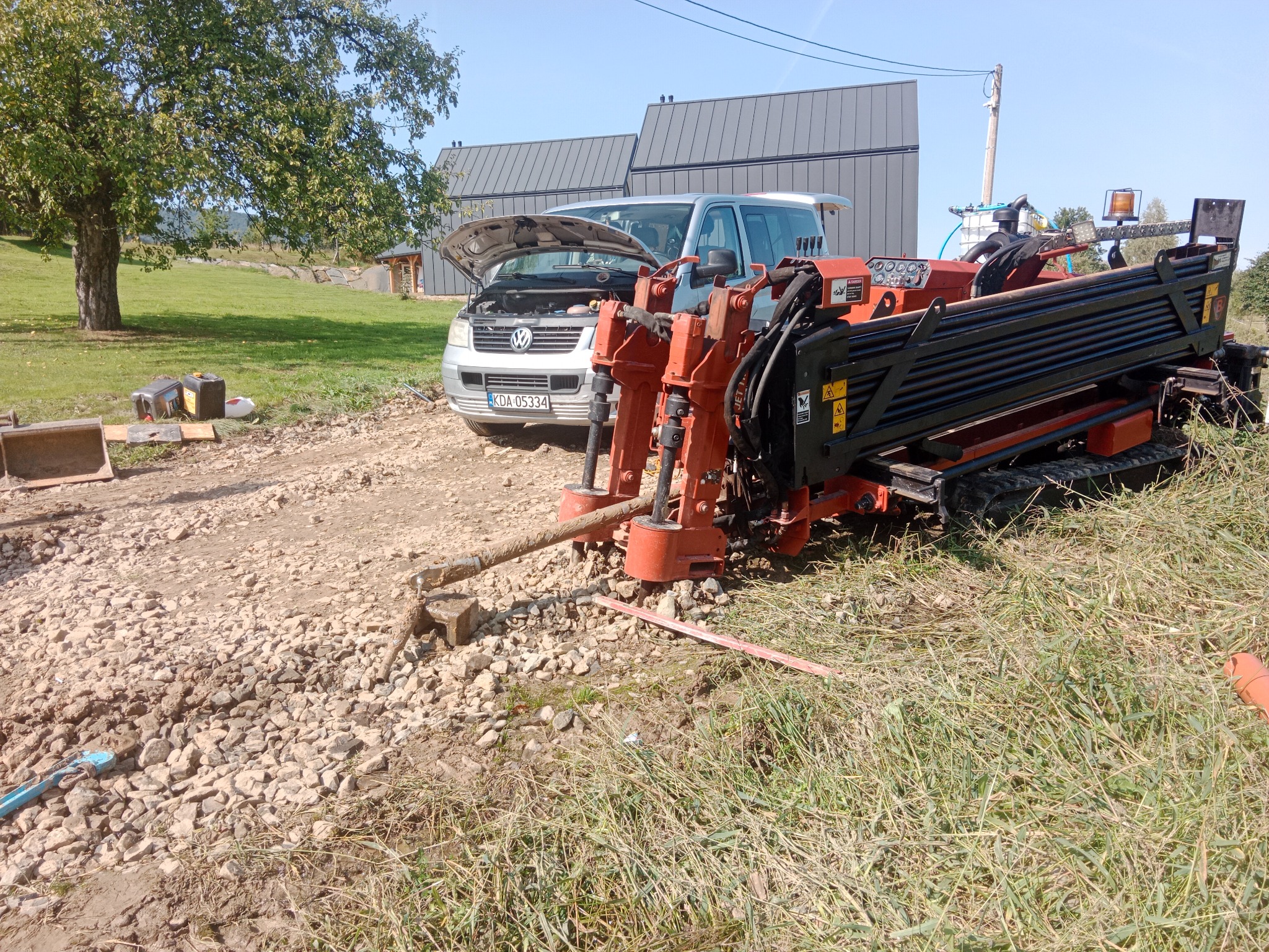 Maszyna do robót ziemnych na żwirowym podłożu, z otwartą maską samochodu w tle. Widoczne elementy instalacji i narzędzia. Dzień słoneczny, teren budowy.