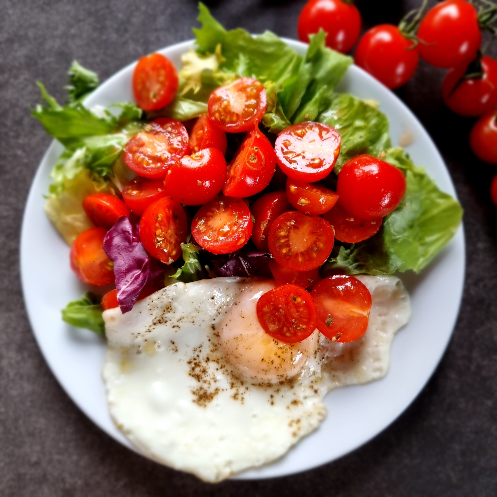 Śniadanie na białym talerzu: sałata, pomidorki koktajlowe i jajko sadzone posypane przyprawami.