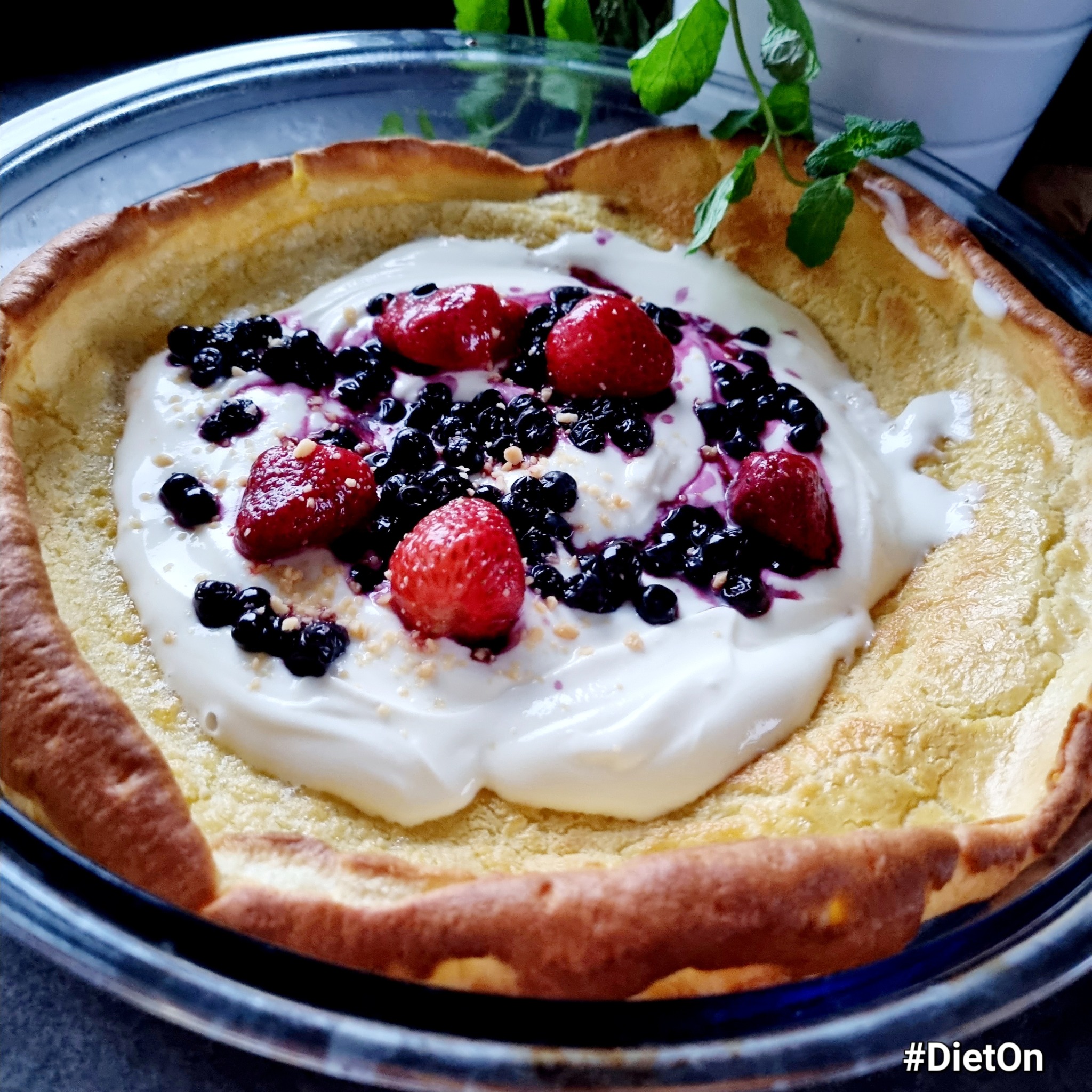 Dutch baby pancake w szklanym naczyniu, udekorowany kremowym jogurtem, świeżymi borówkami, truskawkami i listkami mięty, z widocznym hashtagiem #DietOn.