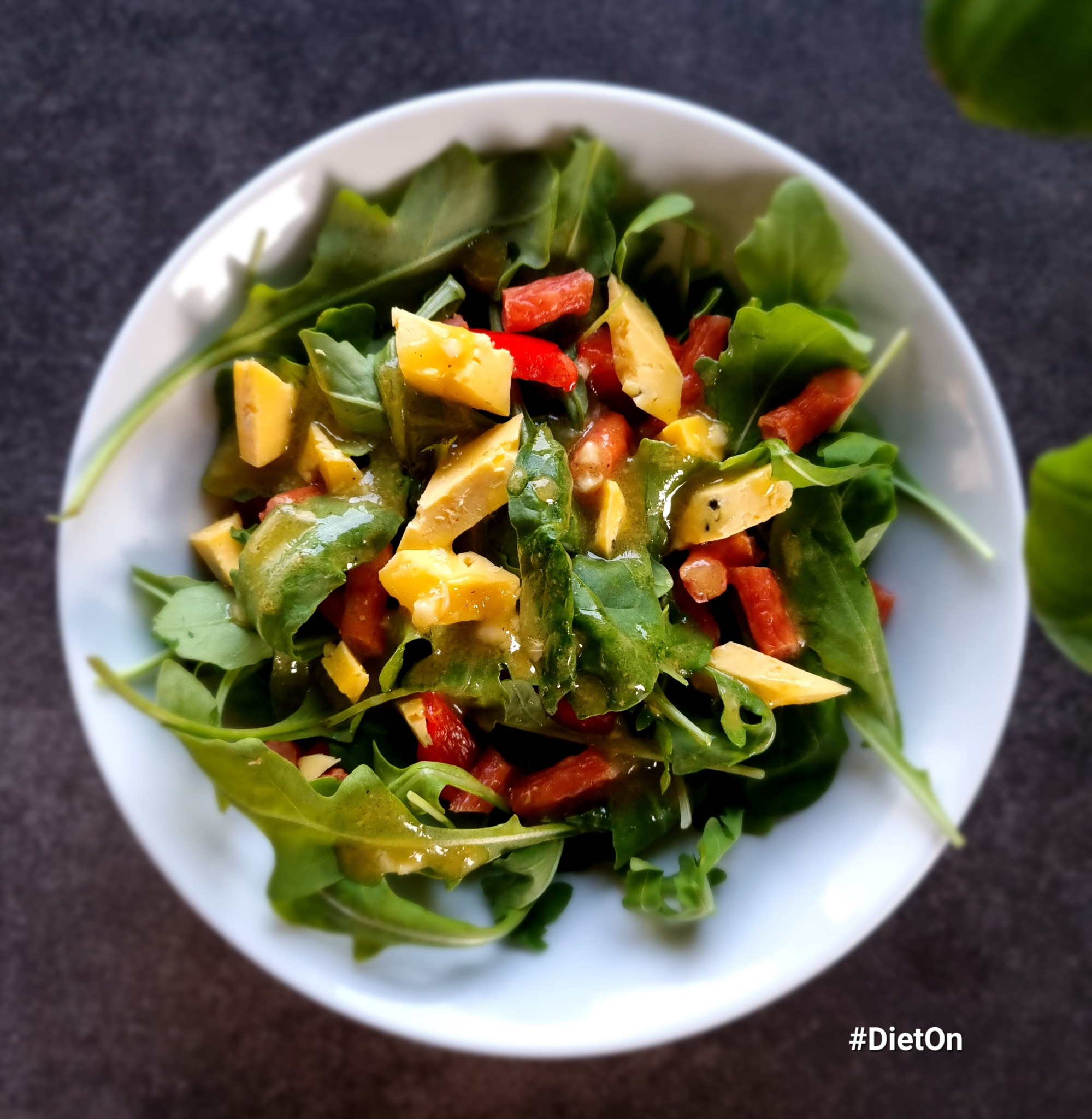 Świeża sałatka z rukoli, papryki i sera w białej misce, polana sosem, widoczna z góry na ciemnym tle.