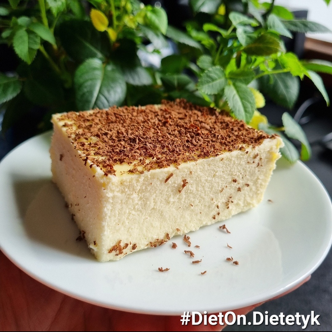 Kwadratowy kawałek sernika na zimno posypany startą czekoladą, na białym talerzu, trzymany w dłoni, z zielonymi roślinami w tle.