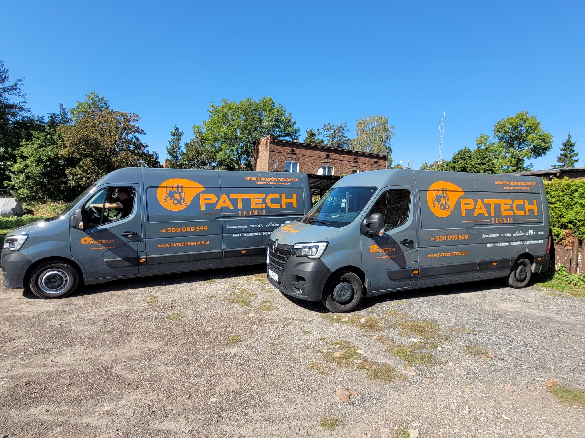Dwa szare samochody dostawcze marki Renault z pomarańczowym logo PATECH na boku, zaparkowane na żwirowym parkingu w słoneczny dzień.