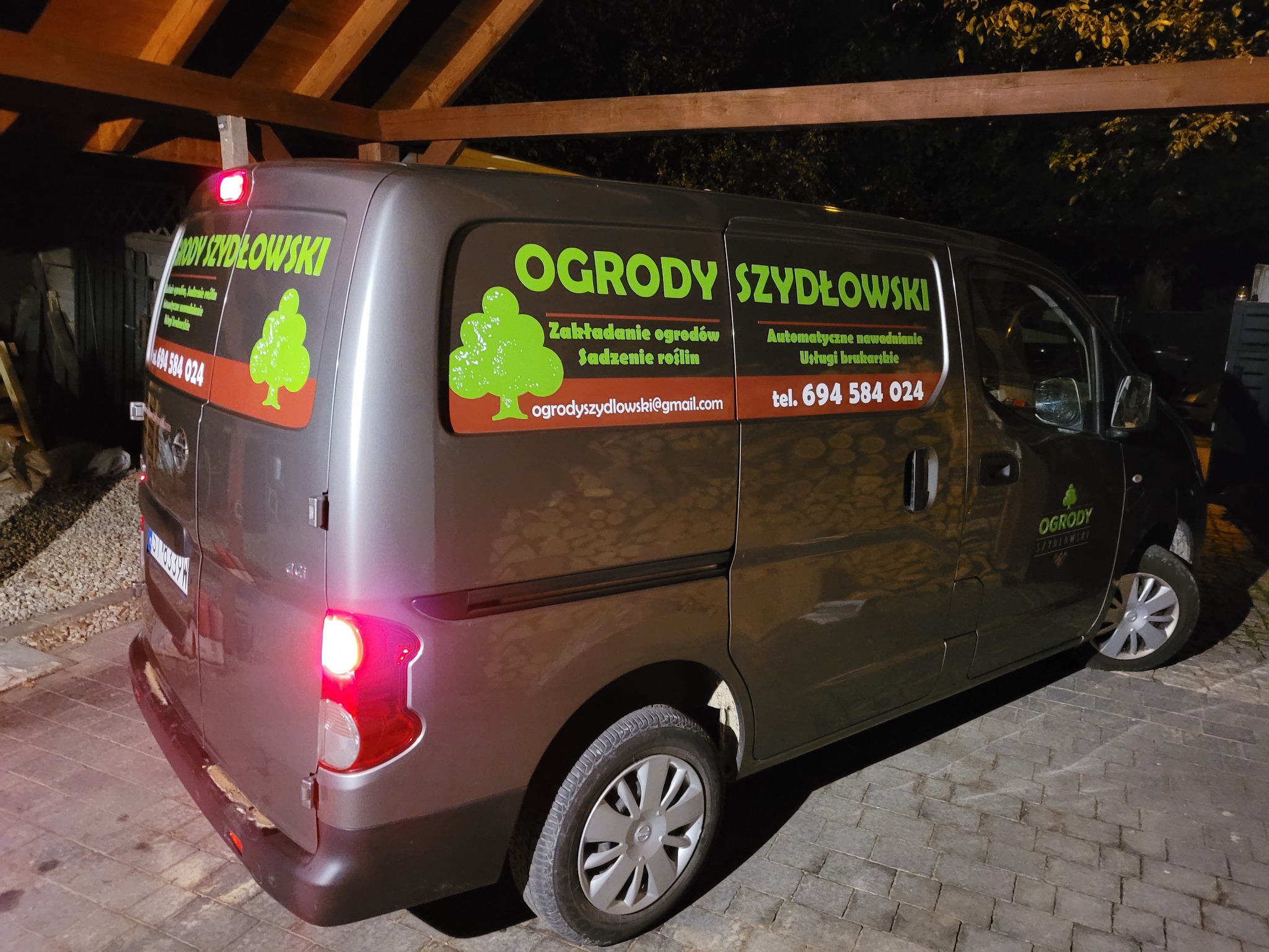 Szary van z reklamą firmy ogrodniczej 'Ogrody Szydłowski', prezentujący logo z zielonym drzewkiem, dane kontaktowe i zakres usług, sfotografowany nocą pod zadaszeniem.