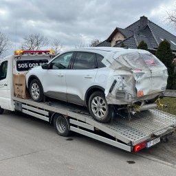 MM Transport Michał Cichosz - Biała laweta z uszkodzonym białym SUV-em na platformie. Widoczny numer telefonu na kabinie lawety i uszkodzony tył pojazdu zabezpieczony folią. Tło: dom i drzewa.