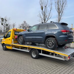 MM Transport Michał Cichosz - Żółta laweta z niebieskim SUV-em na platformie, zabezpieczonym czerwonymi pasami. Widoczne drzewa bez liści i budynek w tle. Ujęcie z boku, na zewnątrz.