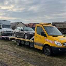 MM Transport Michał Cichosz - Żółta laweta Mercedes z dwoma pojazdami (bus i osobówka) na platformie, na tle pochmurnego nieba i zabudowań. Widoczne oznaczenia firmy i numer telefonu.