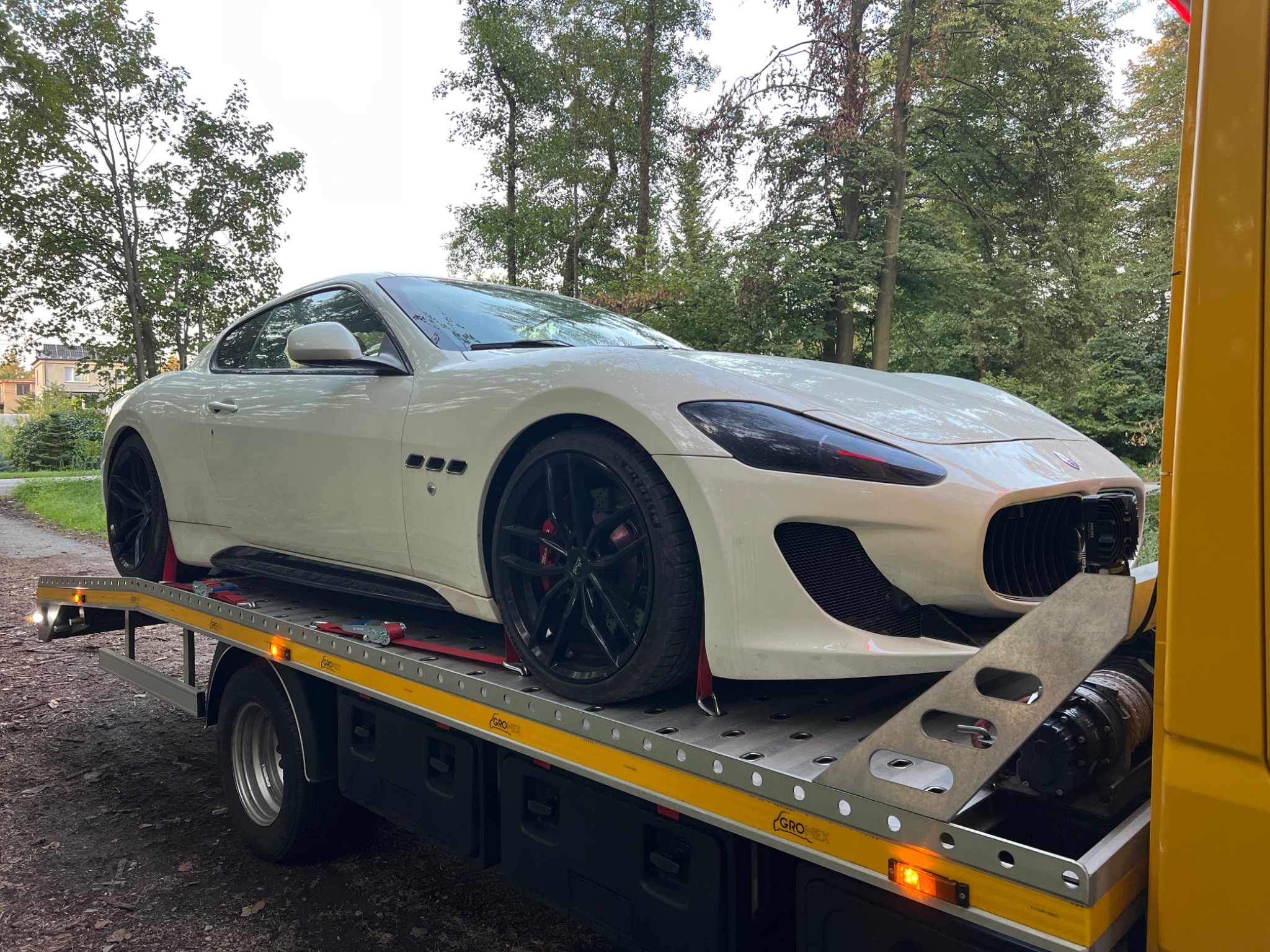Białe sportowe auto Maserati GranTurismo zabezpieczone czerwonymi pasami na żółtej platformie lawety, widok z boku na tle zieleni drzew.