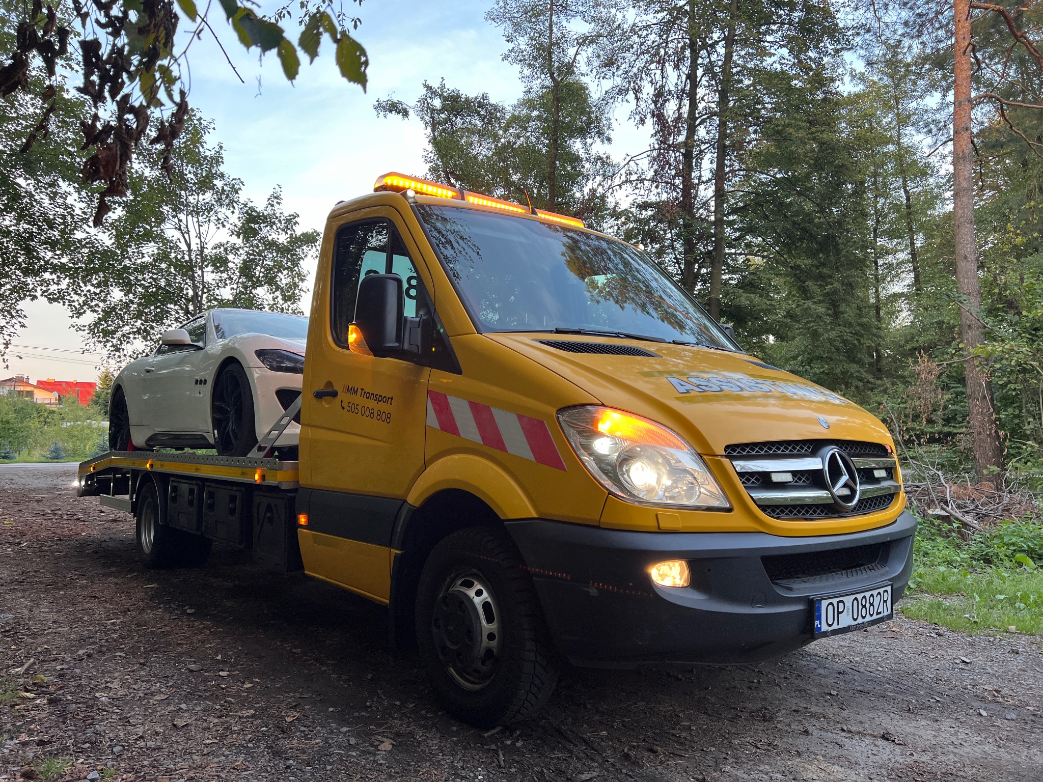 Żółta laweta Mercedes-Benz z białym sportowym samochodem na platformie, zaparkowana na leśnej drodze, w tle drzewa i fragment zabudowań.