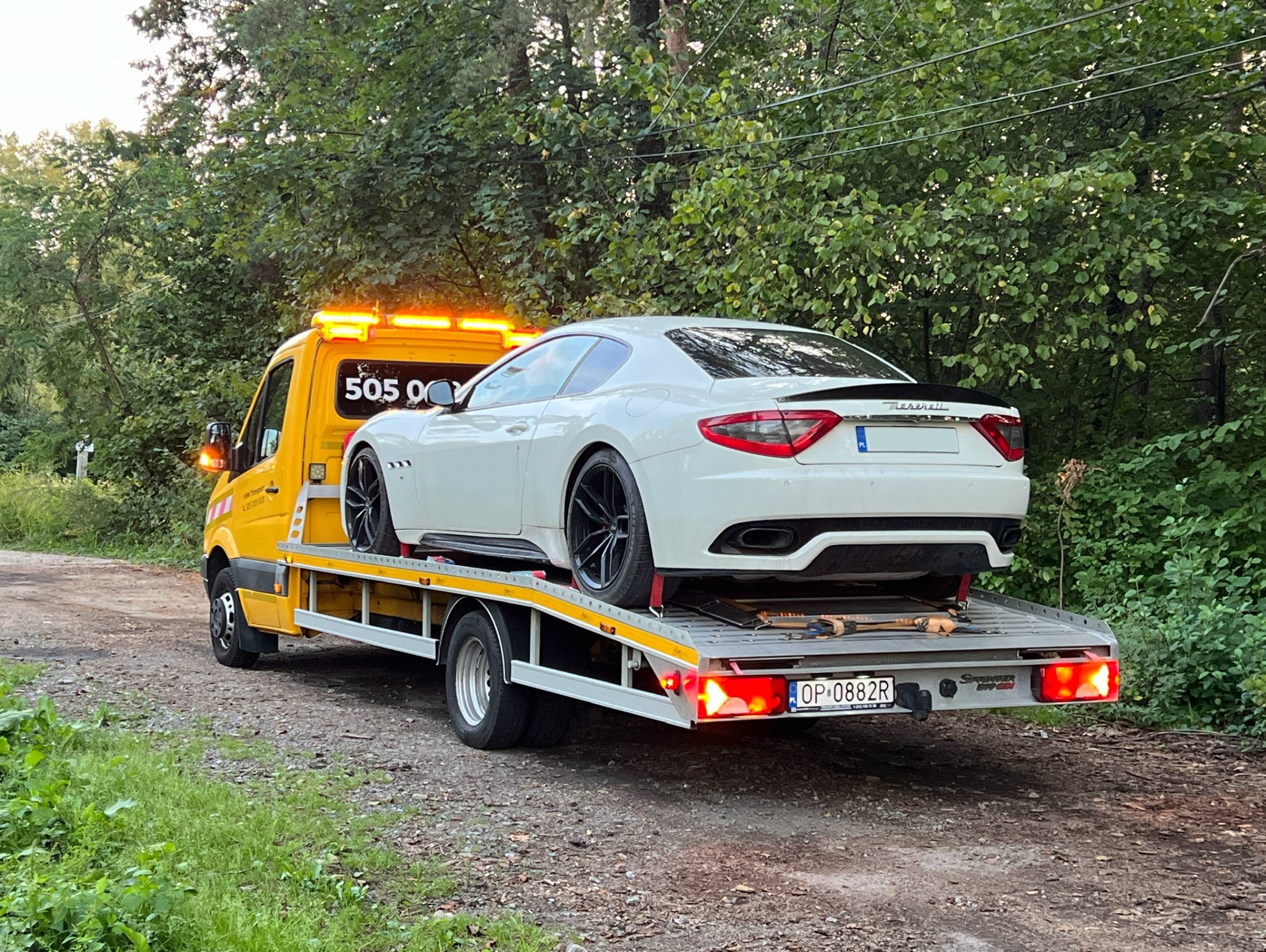 Żółta laweta transportuje białe sportowe auto Maserati GranTurismo z czarnymi felgami na tle zielonego lasu, widoczny numer telefonu na kabinie.