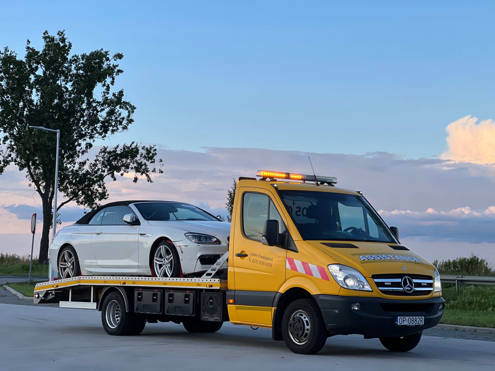 Żółta laweta Mercedes-Benz z włączonymi światłami ostrzegawczymi przewozi uszkodzone białe BMW kabriolet na tle błękitnego nieba o zmierzchu.