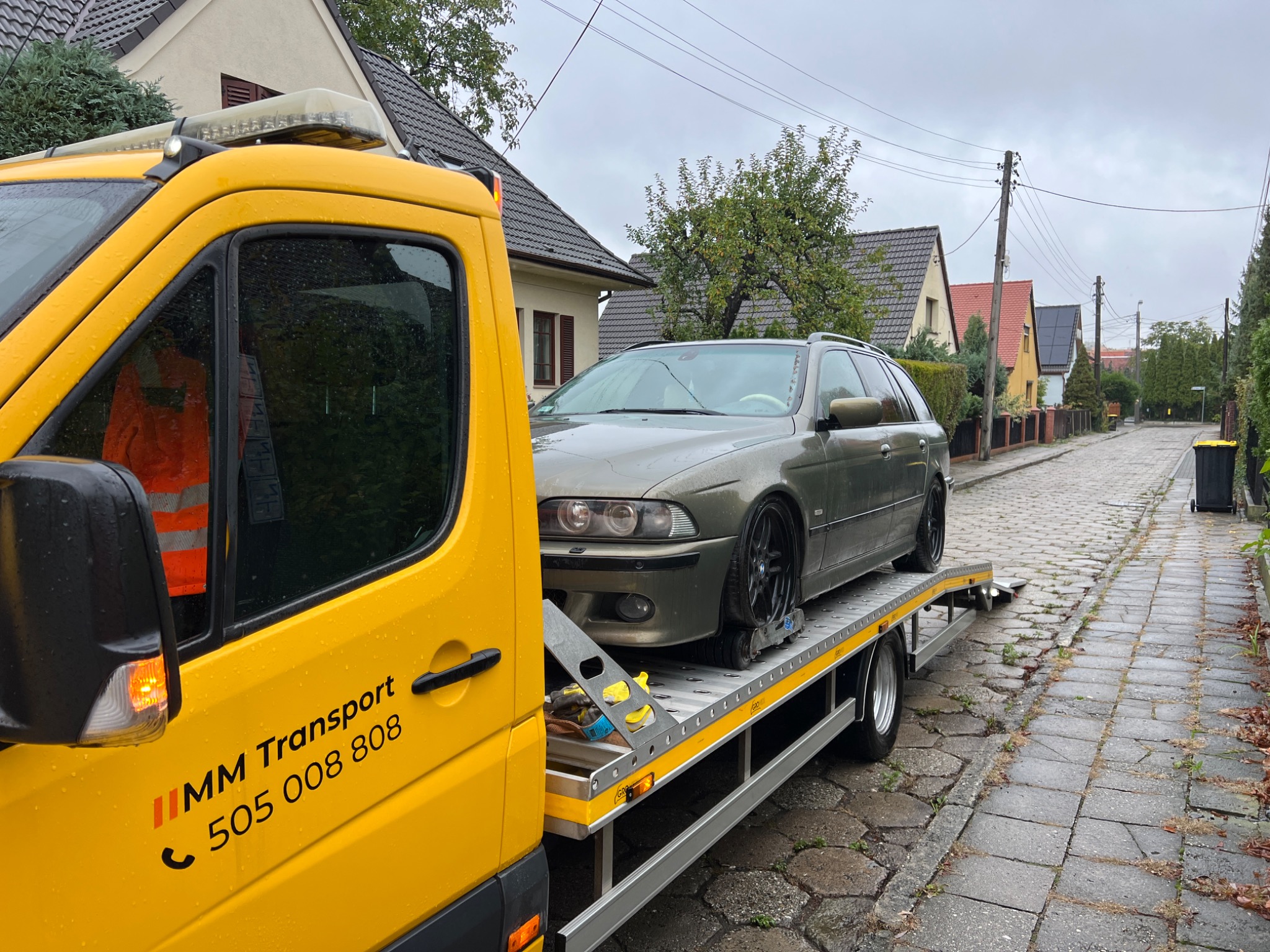 Żółta laweta MM Transport z załadowanym szarym kombi BMW E39 na tle brukowanej ulicy i domów jednorodzinnych w pochmurny dzień.