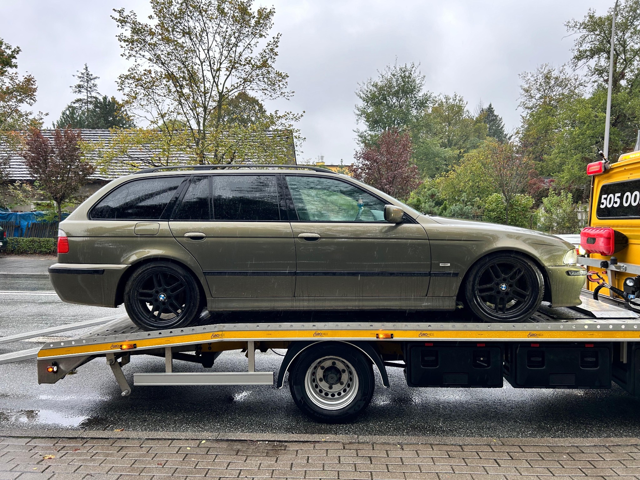 Zielone kombi BMW E39 na żółtej lawecie w pochmurny dzień, widok z boku, mokra nawierzchnia.