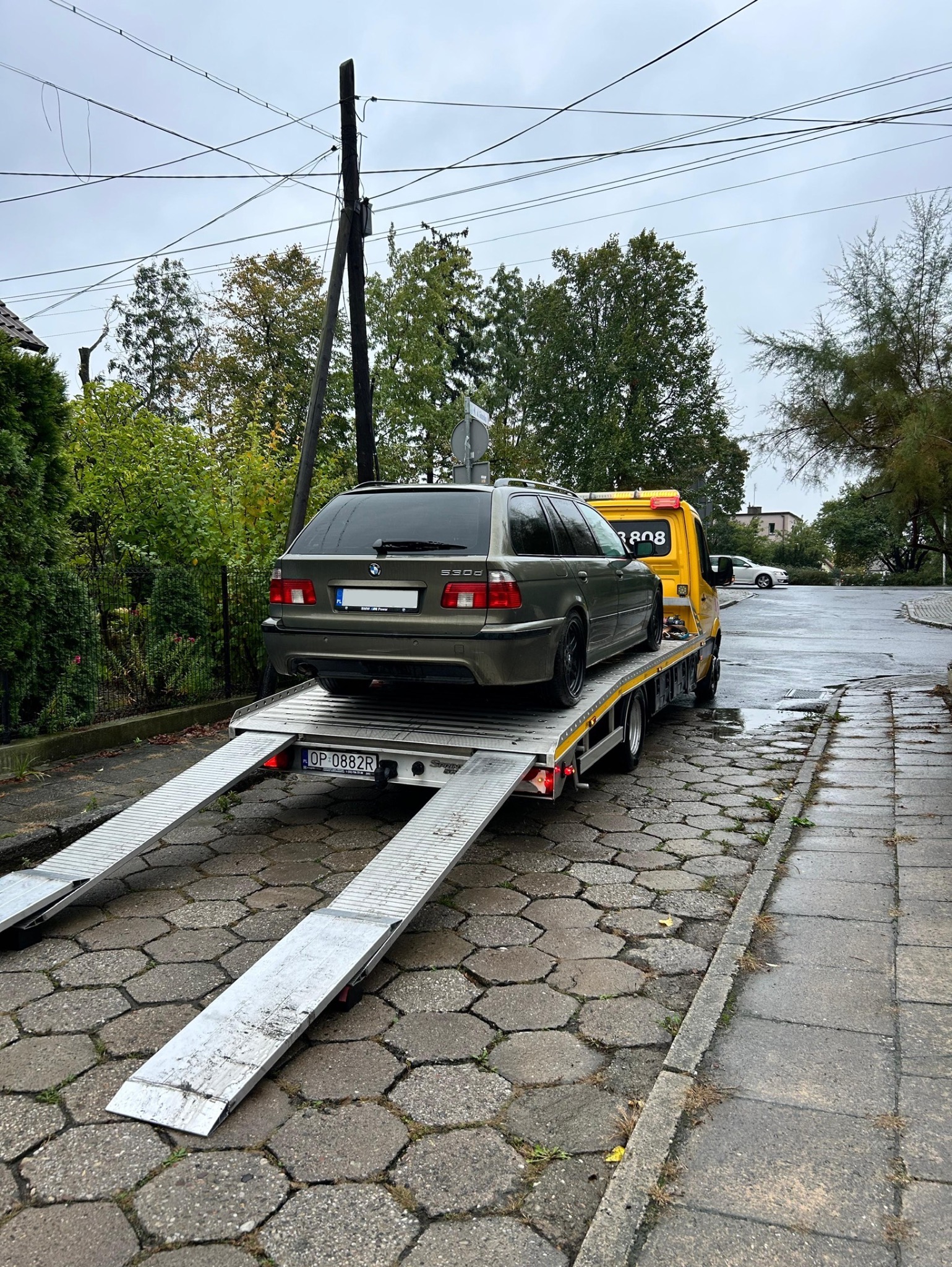 Ciemnozielone kombi BMW serii 5 (E39) na platformie żółtej lawety na brukowanej ulicy w pochmurny dzień. Widoczny numer rejestracyjny lawety i częściowo numer rejestracyjny samochodu.
