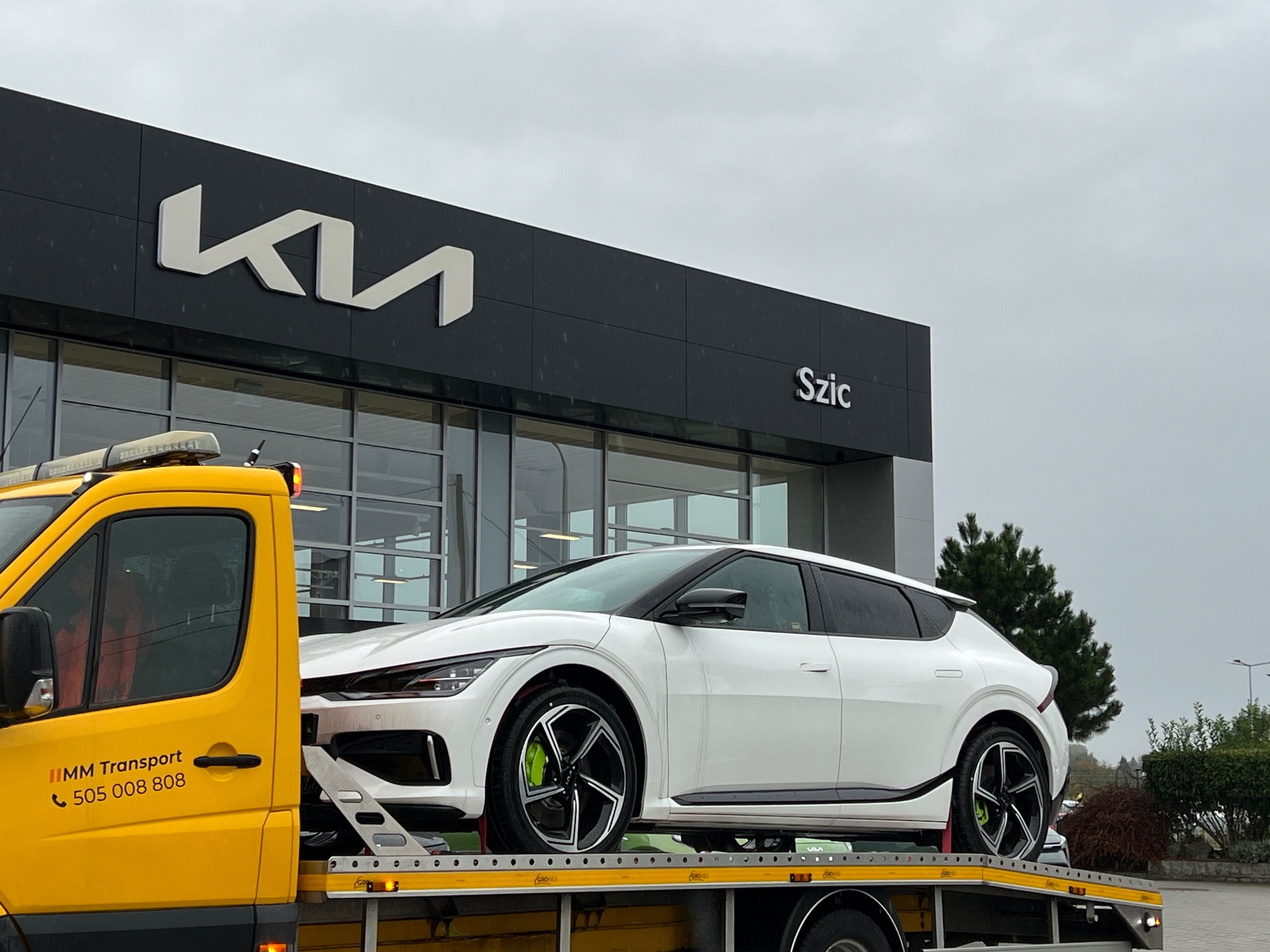 Żółta laweta z białym samochodem elektrycznym Kia EV6 na platformie, przed salonem Kia Szic w pochmurny dzień.
