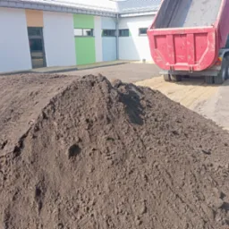 Wyładowany kopiec ciemnej ziemi ogrodowej z wywrotki na przygotowanym podłożu, widoczny budynek z kolorowymi ścianami w tle.