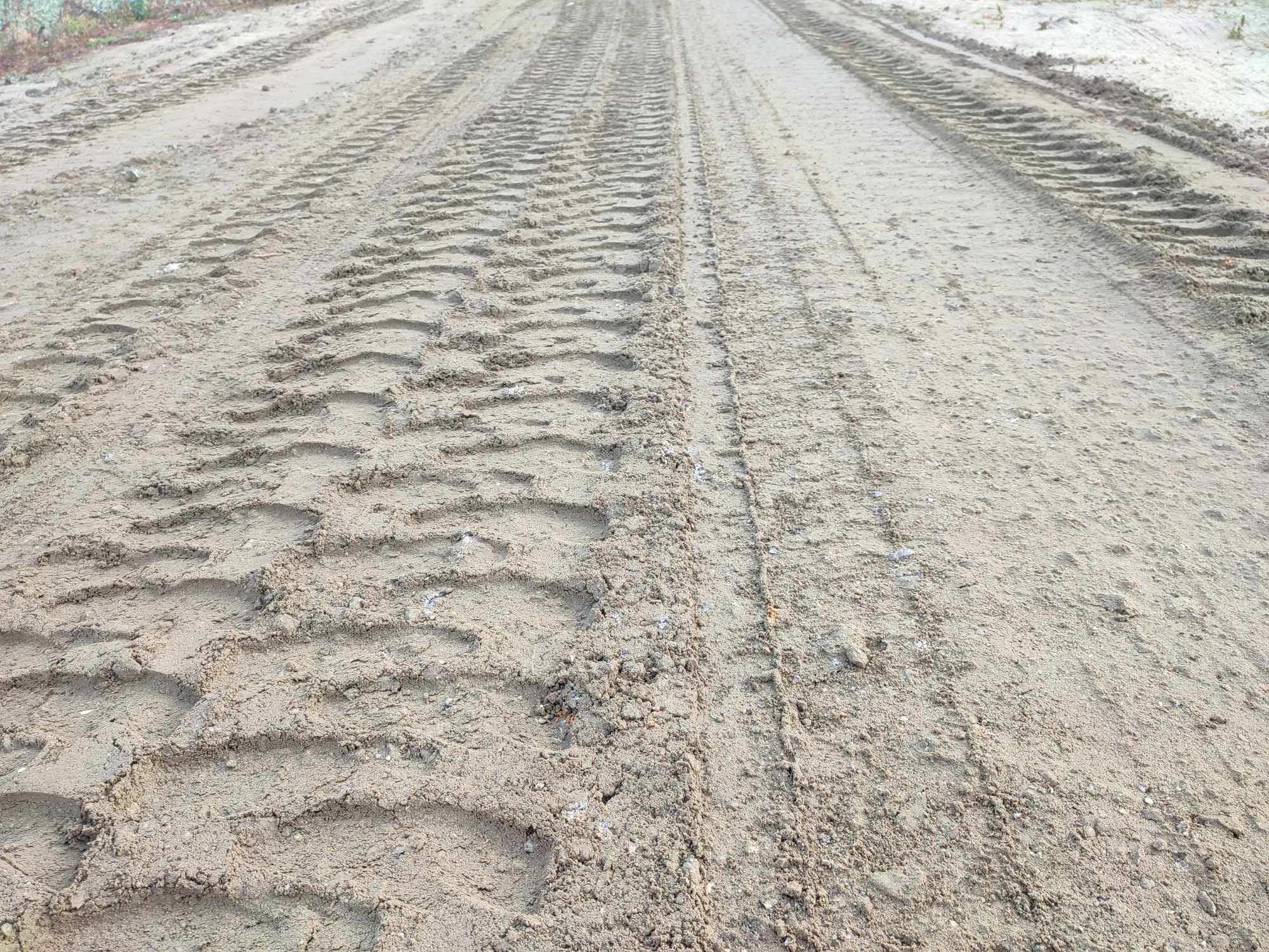 Świeżo utwardzona droga gruntowa z wyraźnymi śladami opon ciężkiego sprzętu budowlanego, widoczne nierówności i drobne grudki ziemi.