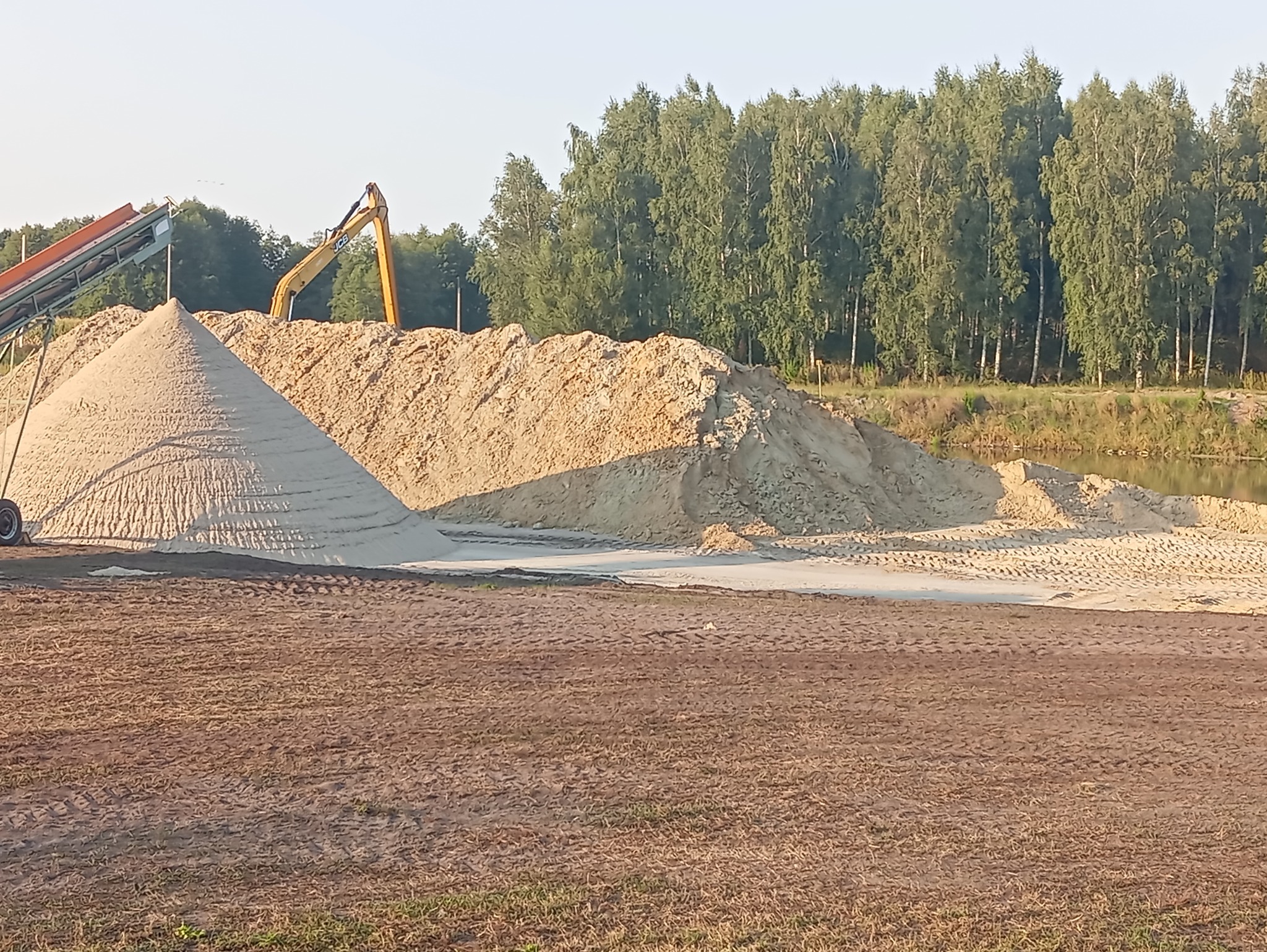 Kopalnia piasku z kopcami usypanego materiału, taśmociągiem i koparką JCB w tle, obok zbiornika wodnego i lasu.