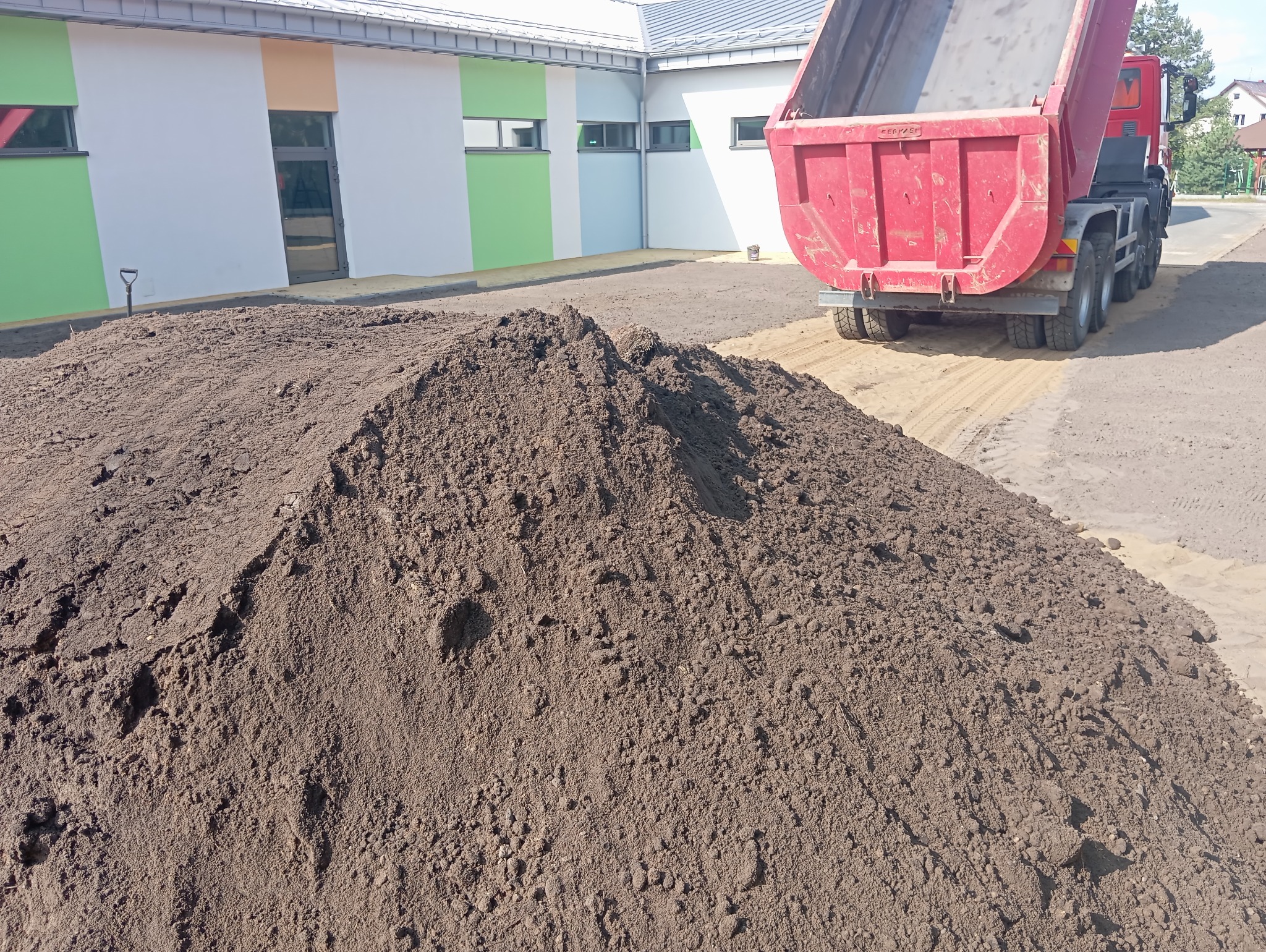 Wyładowany kopiec ciemnej ziemi ogrodowej z wywrotki na przygotowanym podłożu, widoczny budynek z kolorowymi ścianami w tle.